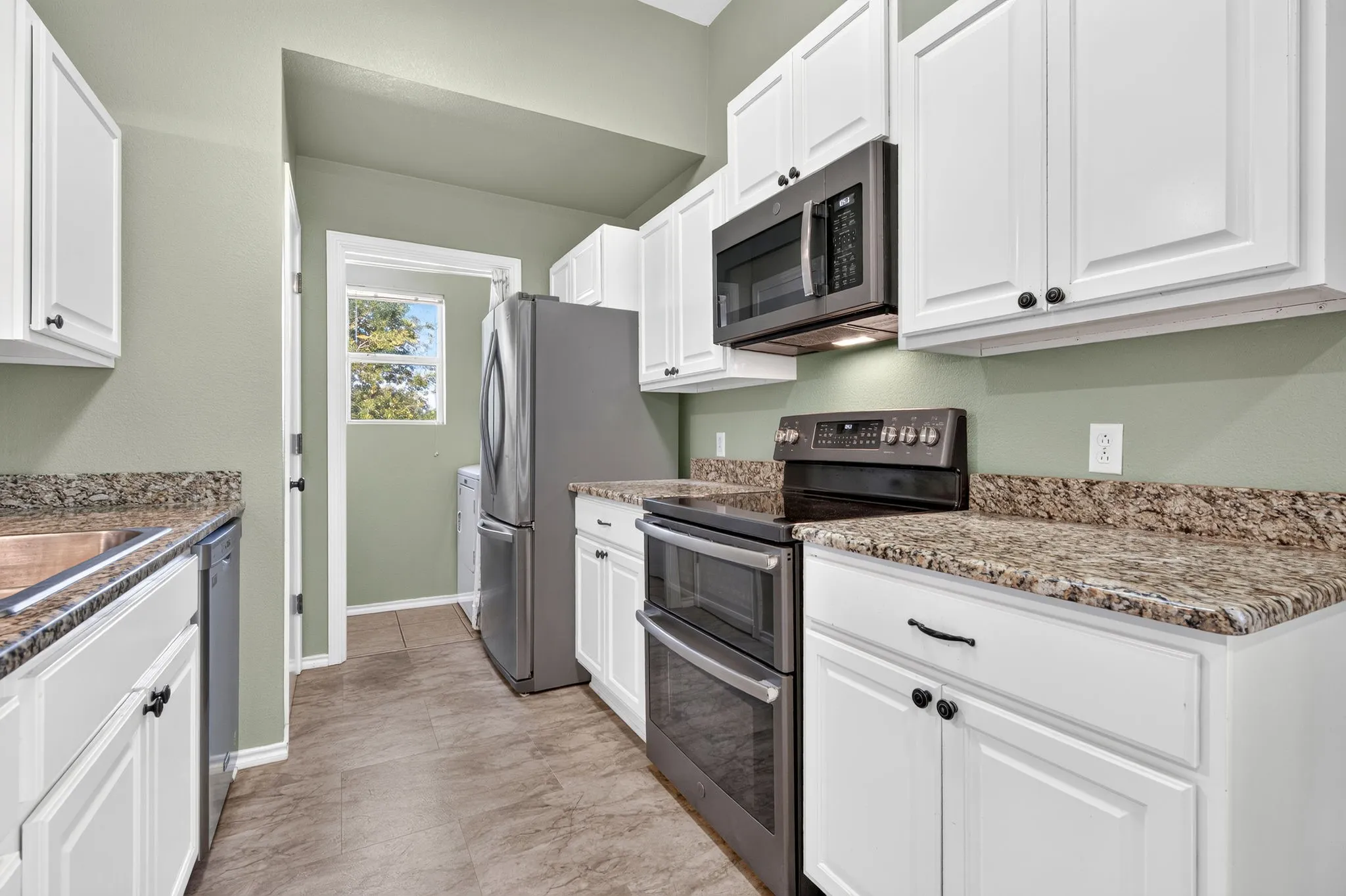 White shaker-style cabinetry, granite countertops, and stainless steel appliances.  View into laundry room.
