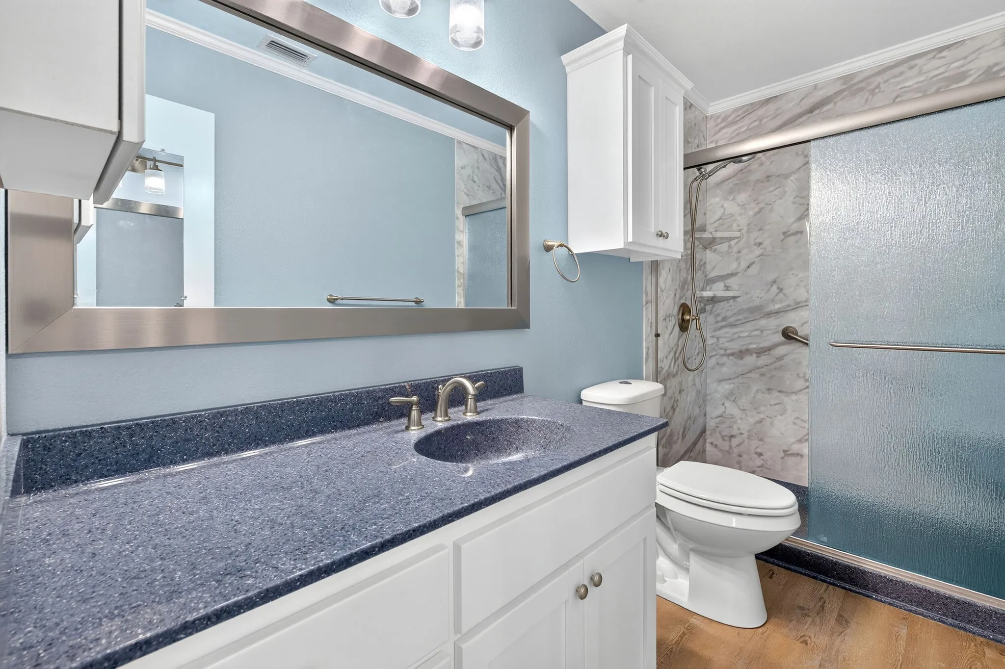 Vanity with dark speckled countertop and sink, white cabinetry, brushed nickel faucet, large framed mirror, and a shower with sliding glass door.