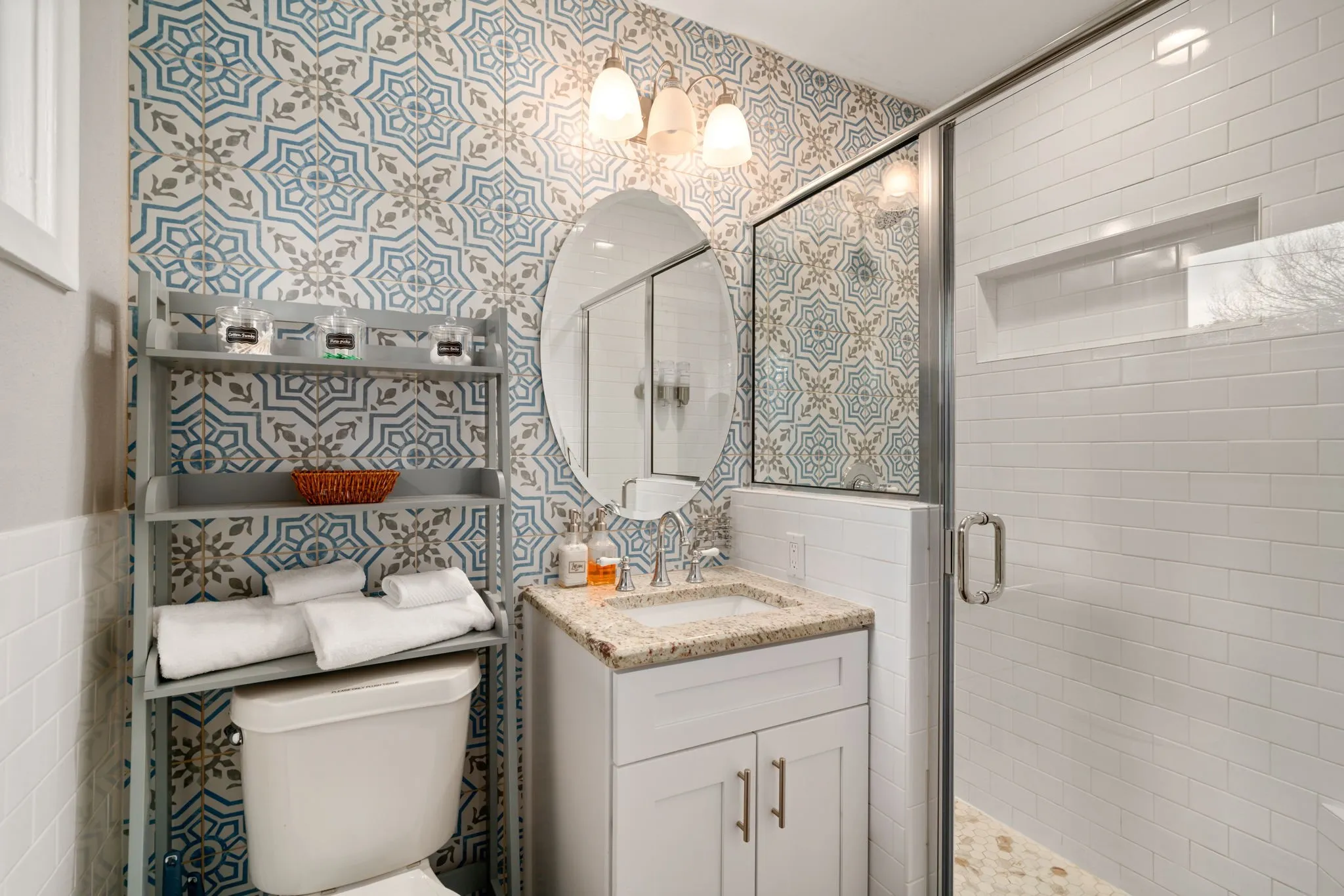 Custom patterned wall tile, vanity with stone countertop, oval mirror, walk-in shower with glass enclosure, white subway tile shower surround