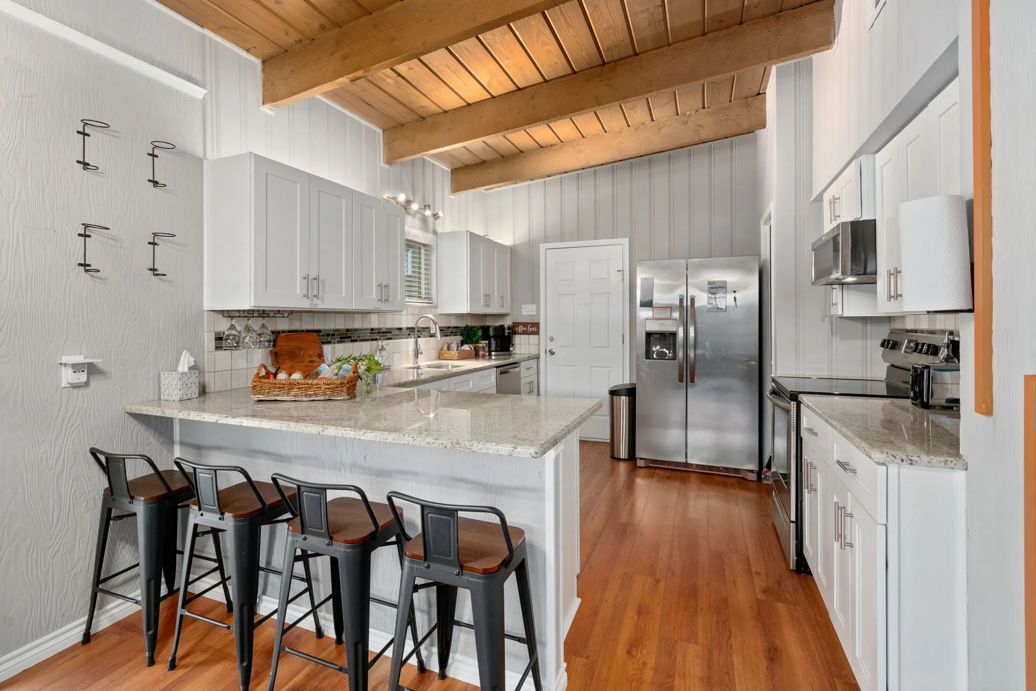Vaulted wood-beamed ceiling, white shaker cabinetry, stainless steel appliances, light-toned granite countertops, and wood-finish flooring