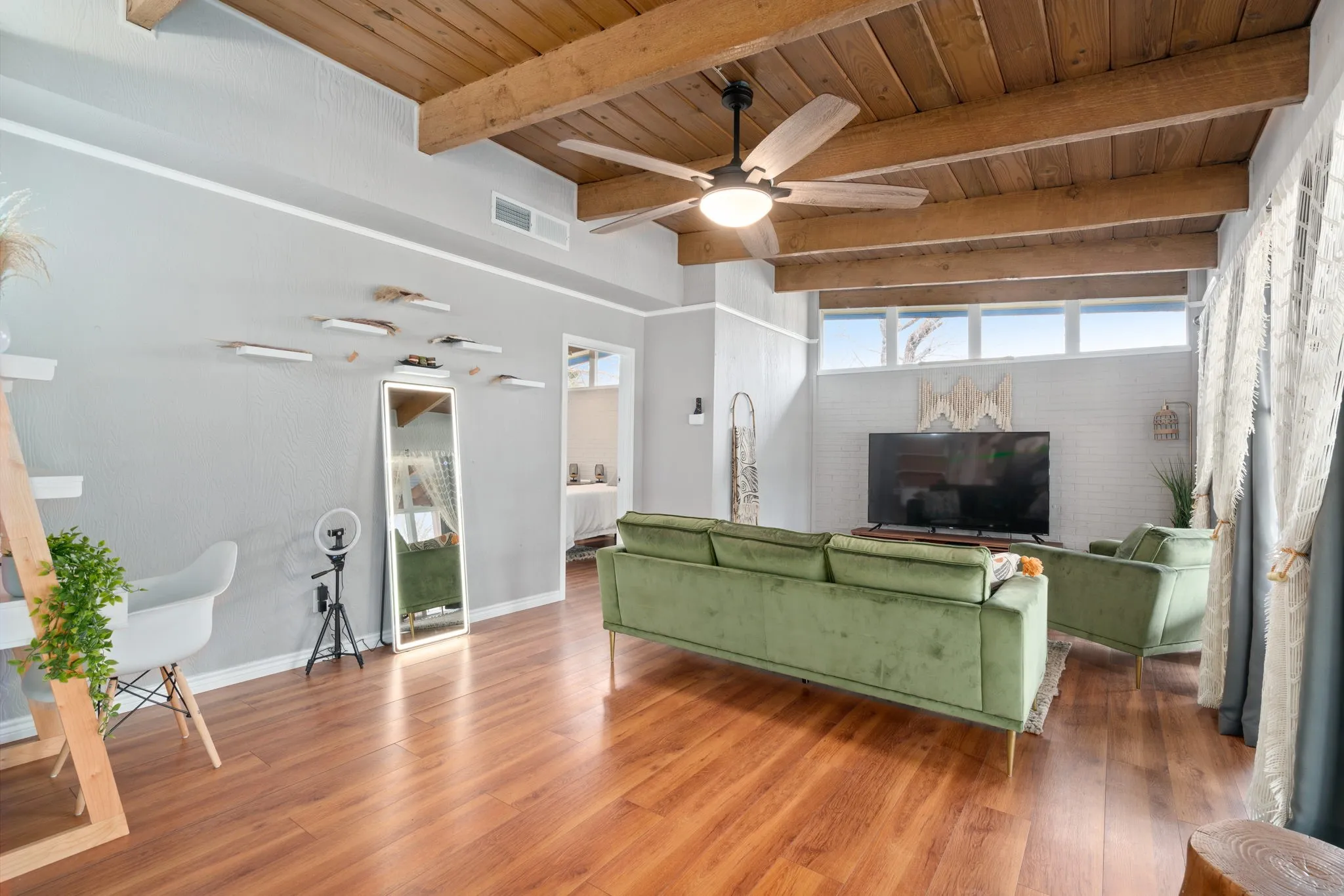 Wood-finish flooring, exposed wood beam ceiling, contemporary ceiling fan with integrated lighting, multiple transom windows, and white brick accent wall