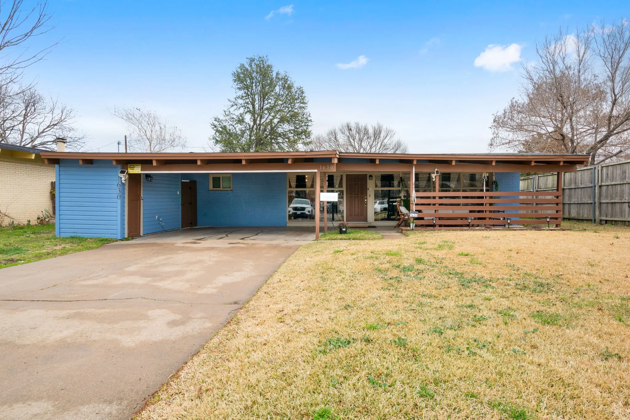 Single-story structure with blue siding, a covered carport, and a front patio with a wood slatted fence