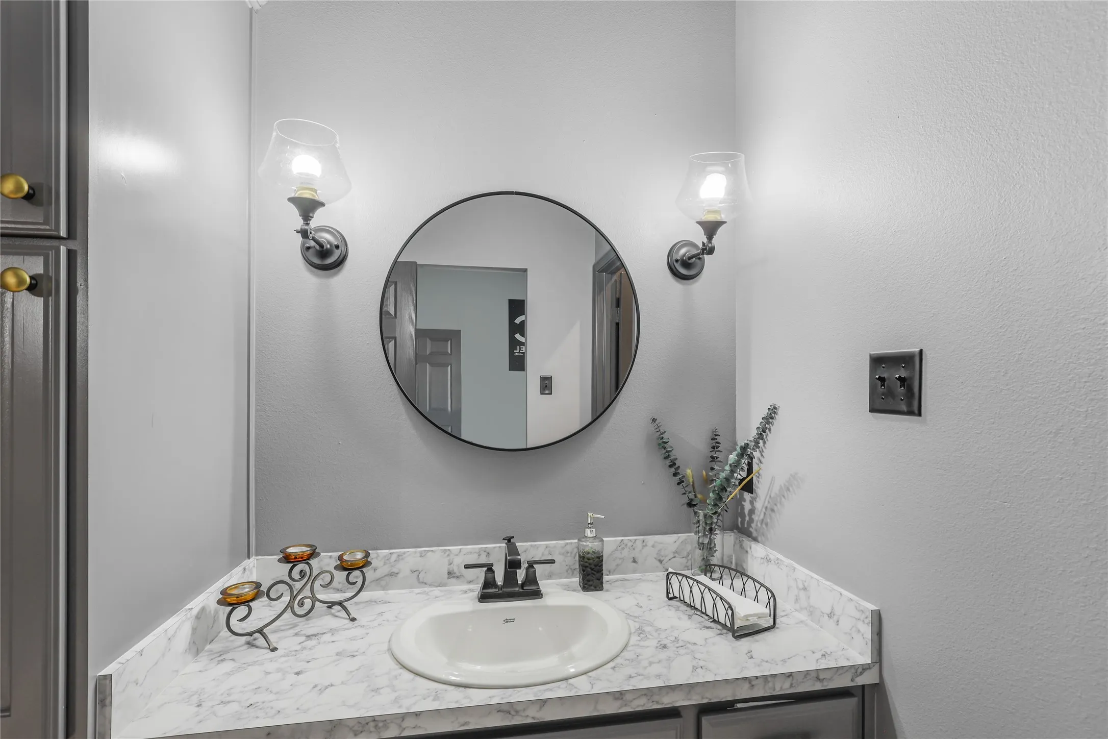 Built-in vanity with a circular mirror, two wall-mounted light fixtures, an integrated sink, and gray cabinetry with gold-toned hardware
