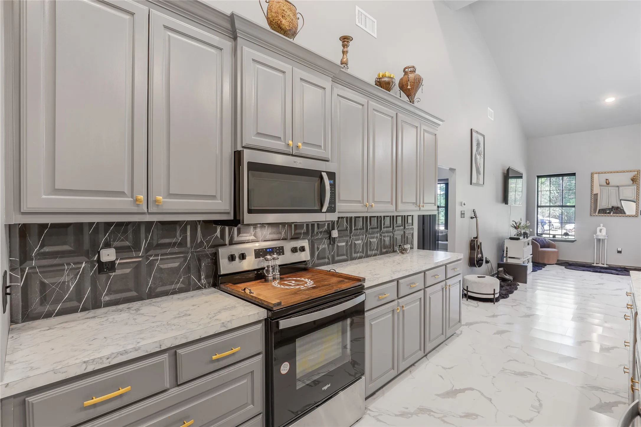 Gray cabinetry with gold-tone hardware, stainless steel appliances, patterned backsplash, light-toned countertops, and vaulted ceilings