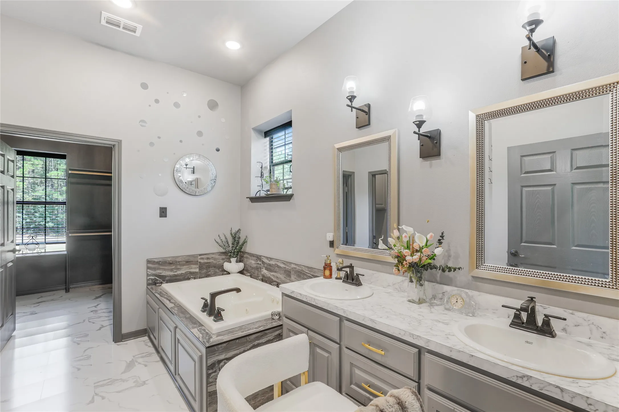 Double vanity with undermount sinks, wall-mounted light fixtures, framed mirrors, soaking tub with dark bronze fixtures, and grey wood-finish cabinetry