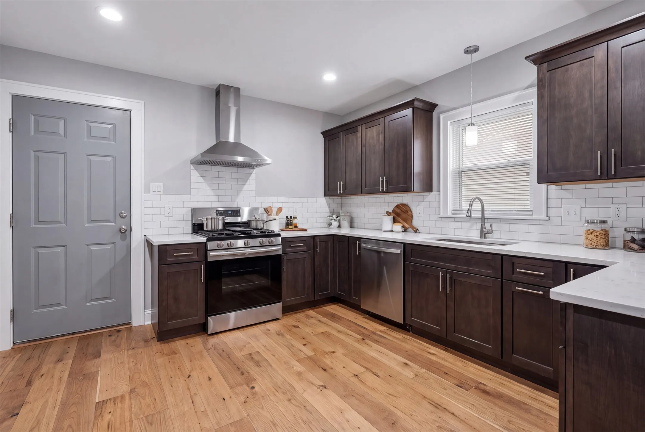 Virtually Altered. Kitchen featuring wood-finish flooring, dark wood cabinetry, white subway tile backsplash, stainless steel appliances, and light-toned countertops
