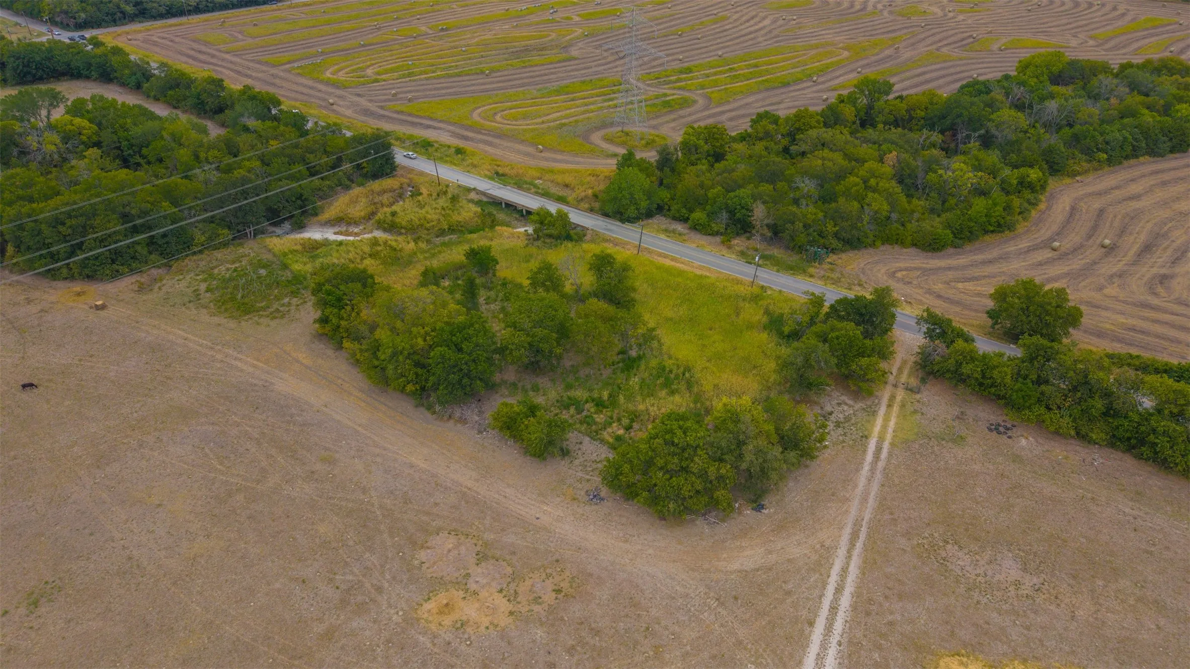 Expansive agricultural land featuring fields with visible crop patterns, a paved road, natural tree lines, and a visible power line infrastructure