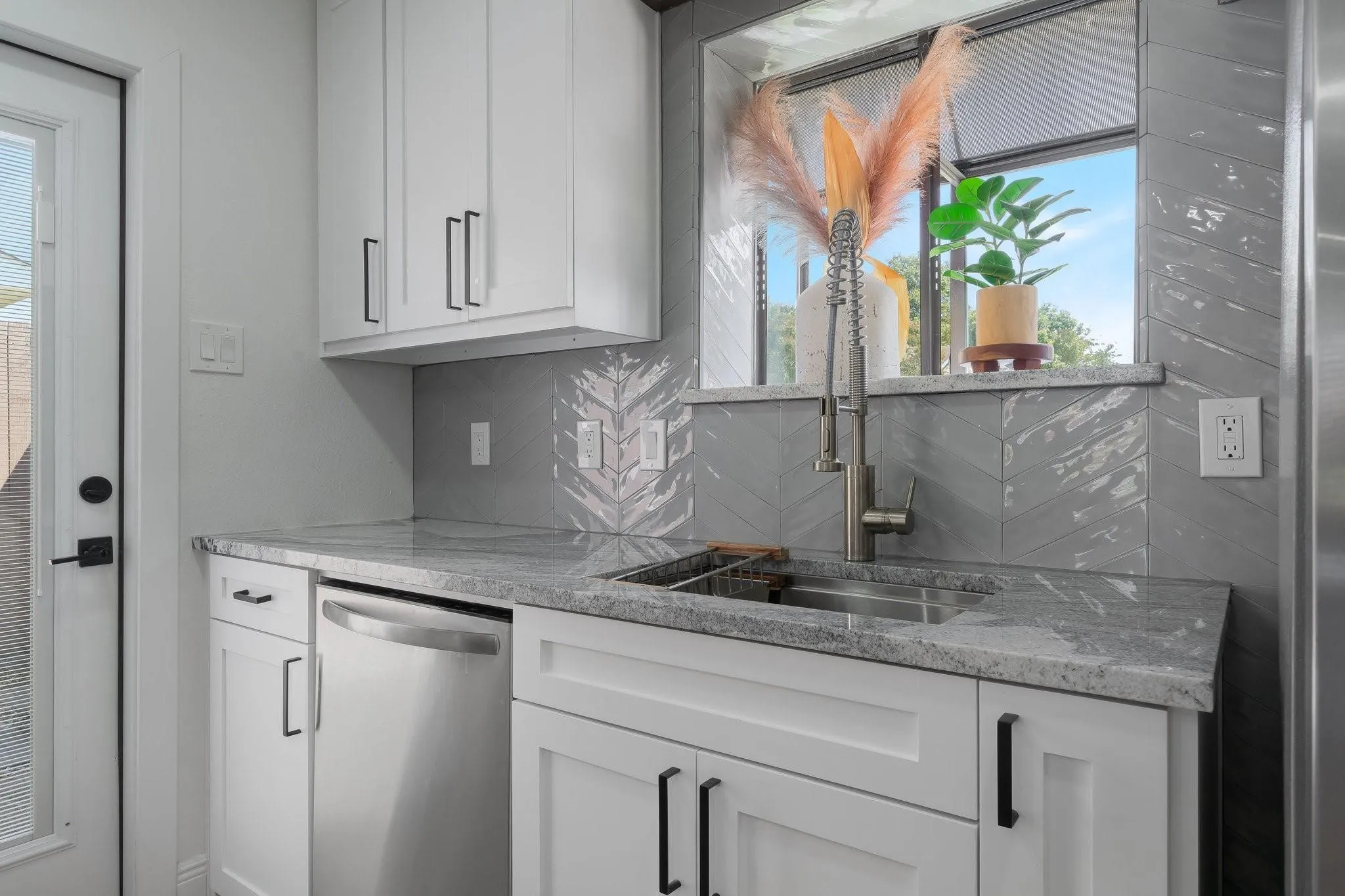 Modern kitchen featuring white shaker-style cabinetry, stainless steel appliances including a dishwasher, gray tile backsplash with geometric pattern detailing, quartz countertops, and a professional-grade pull-down faucet