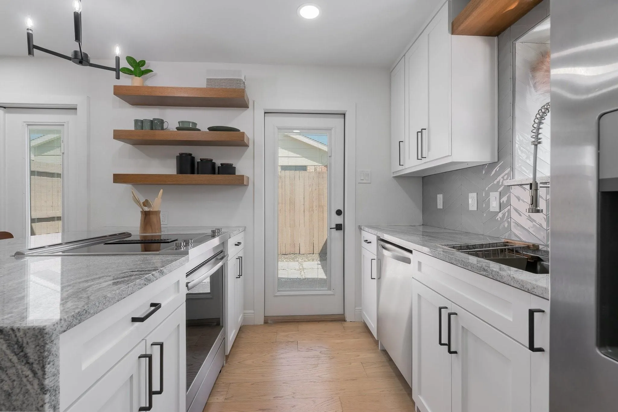 White cabinetry with black hardware, stone countertops, wood-finish open shelving, built-in stainless steel appliances, and a tile backsplash