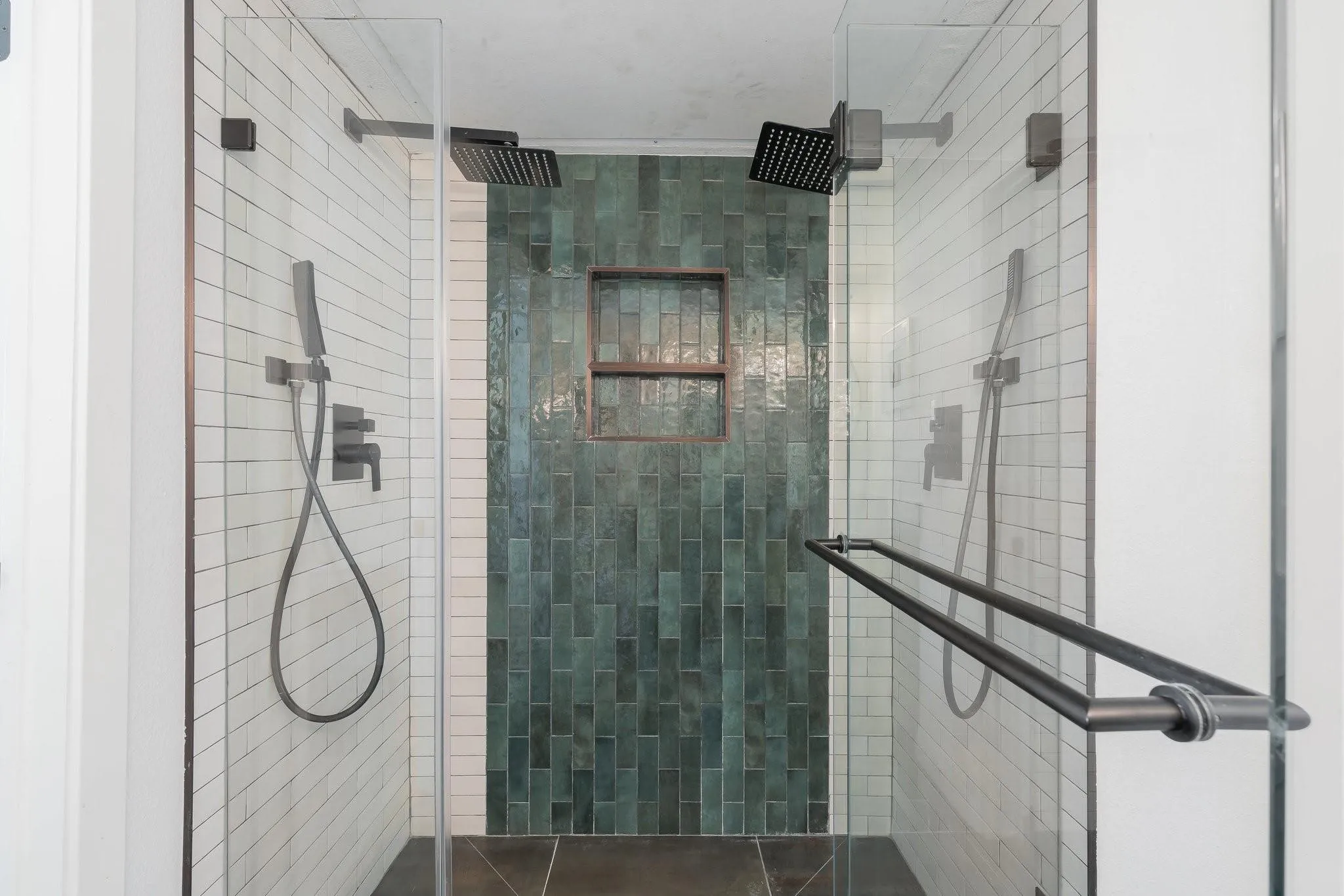 Glass-enclosed shower featuring dual rain shower heads, a handheld shower fixture, a recessed tiled niche, and dark-toned floor tiling