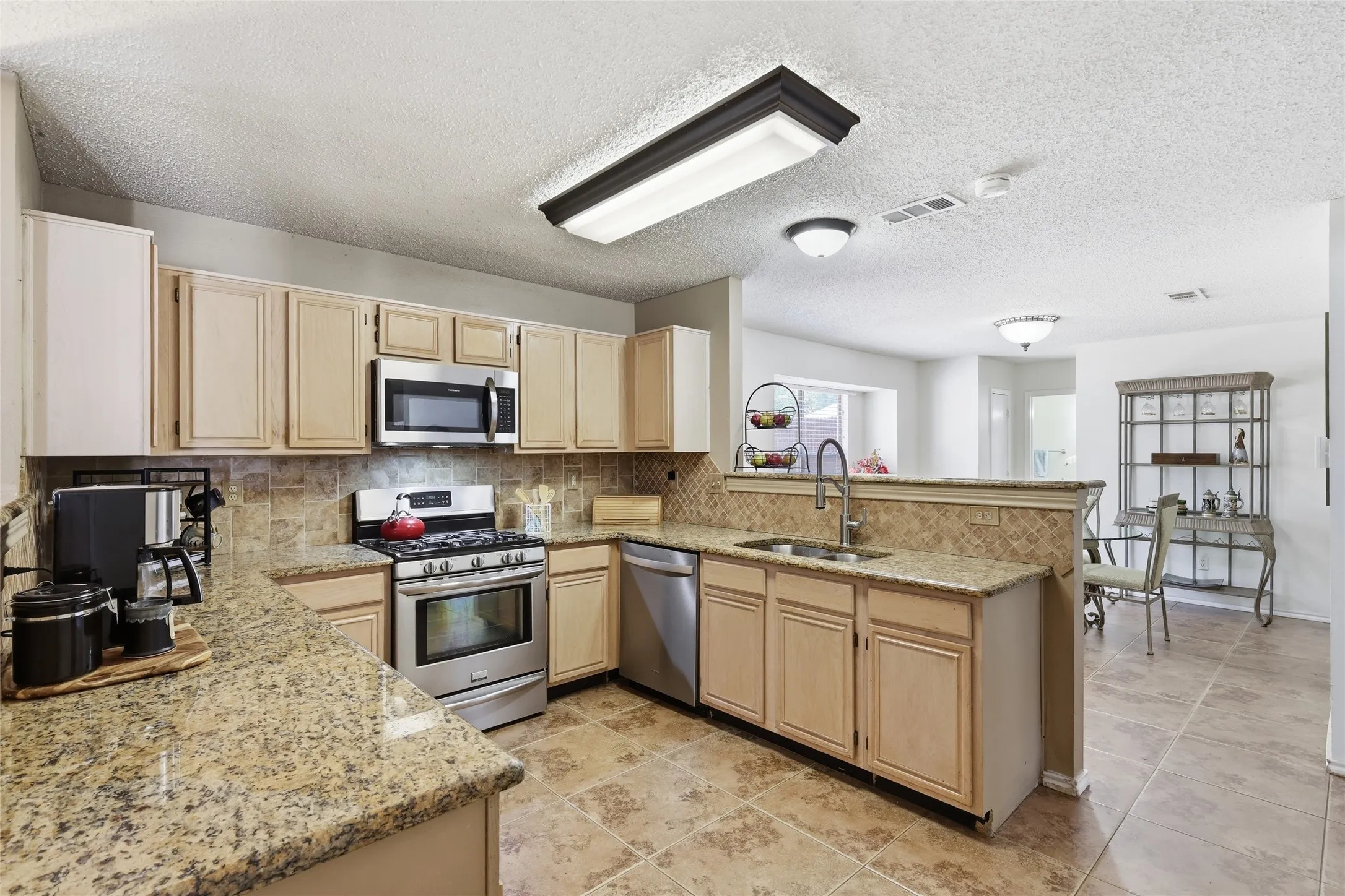 Kitchen featuring granite-look countertops, stainless steel appliances, a tiled backsplash, wood-finish cabinetry, and tiled flooring