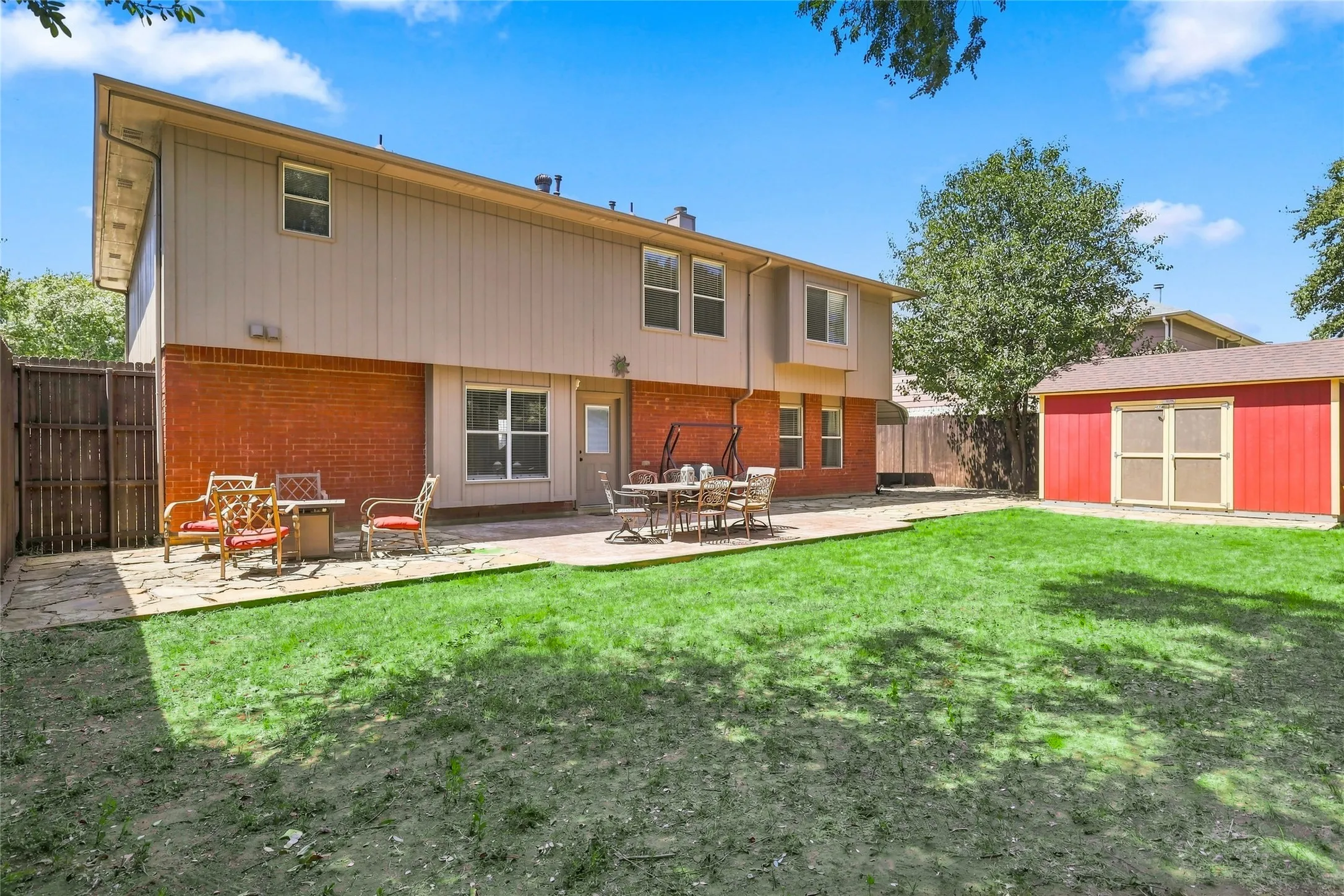 Expansive backyard featuring a red brick and beige panel exterior, a large stone patio, a green lawn, and a red storage shed