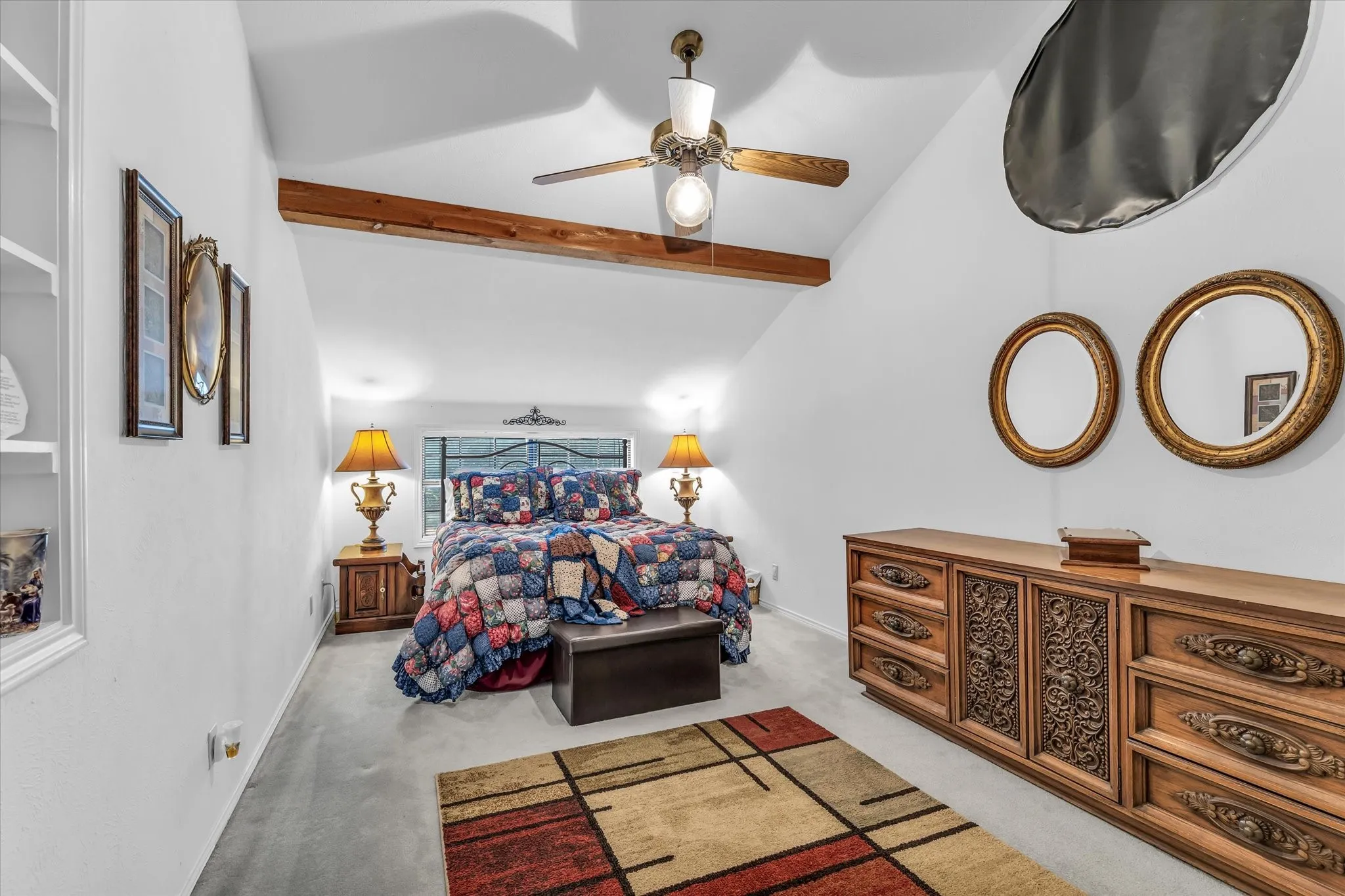 Vaulted ceiling with exposed wood beam, ceiling fan with integrated lighting, built-in shelving, carpet flooring, and a large window