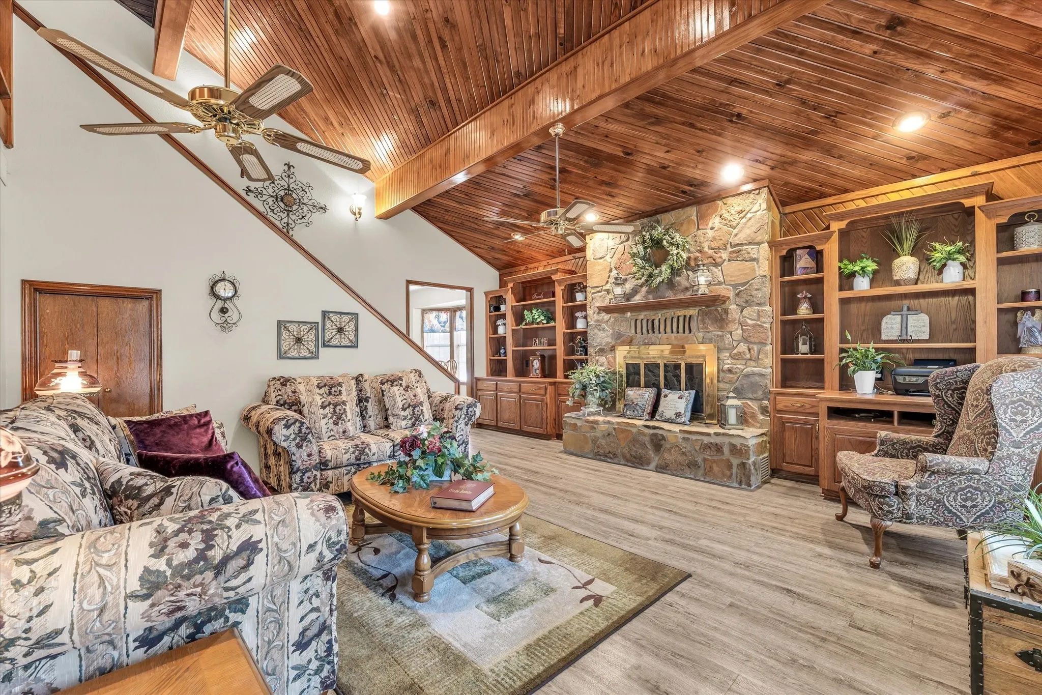 Vaulted wood-paneled ceiling, stone fireplace with brass accents, built-in shelving and cabinetry, wood-finish flooring, and a ceiling fan
