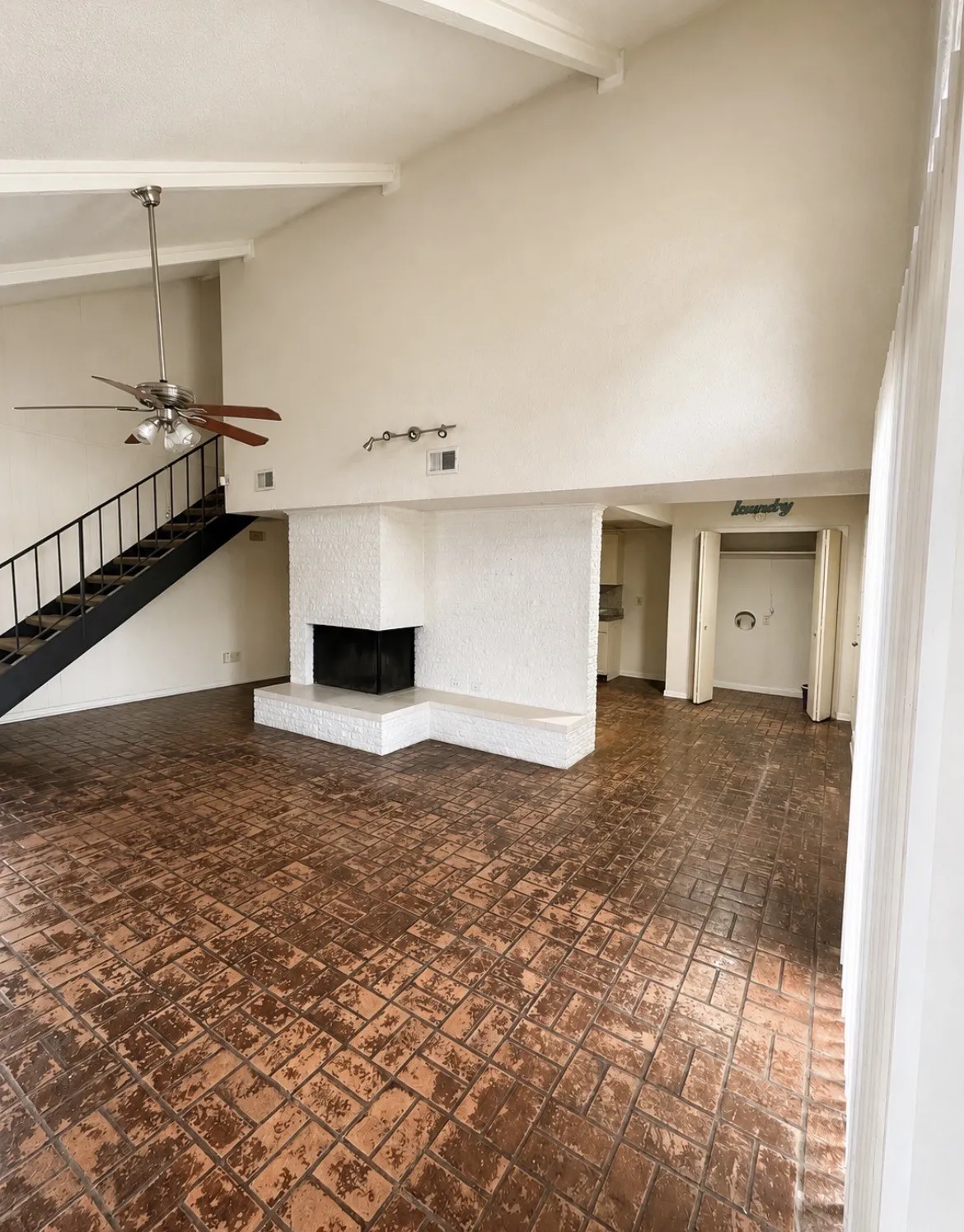Vaulted ceilings with exposed beams, brick flooring, a corner fireplace, a ceiling fan, and an open staircase with metal railing