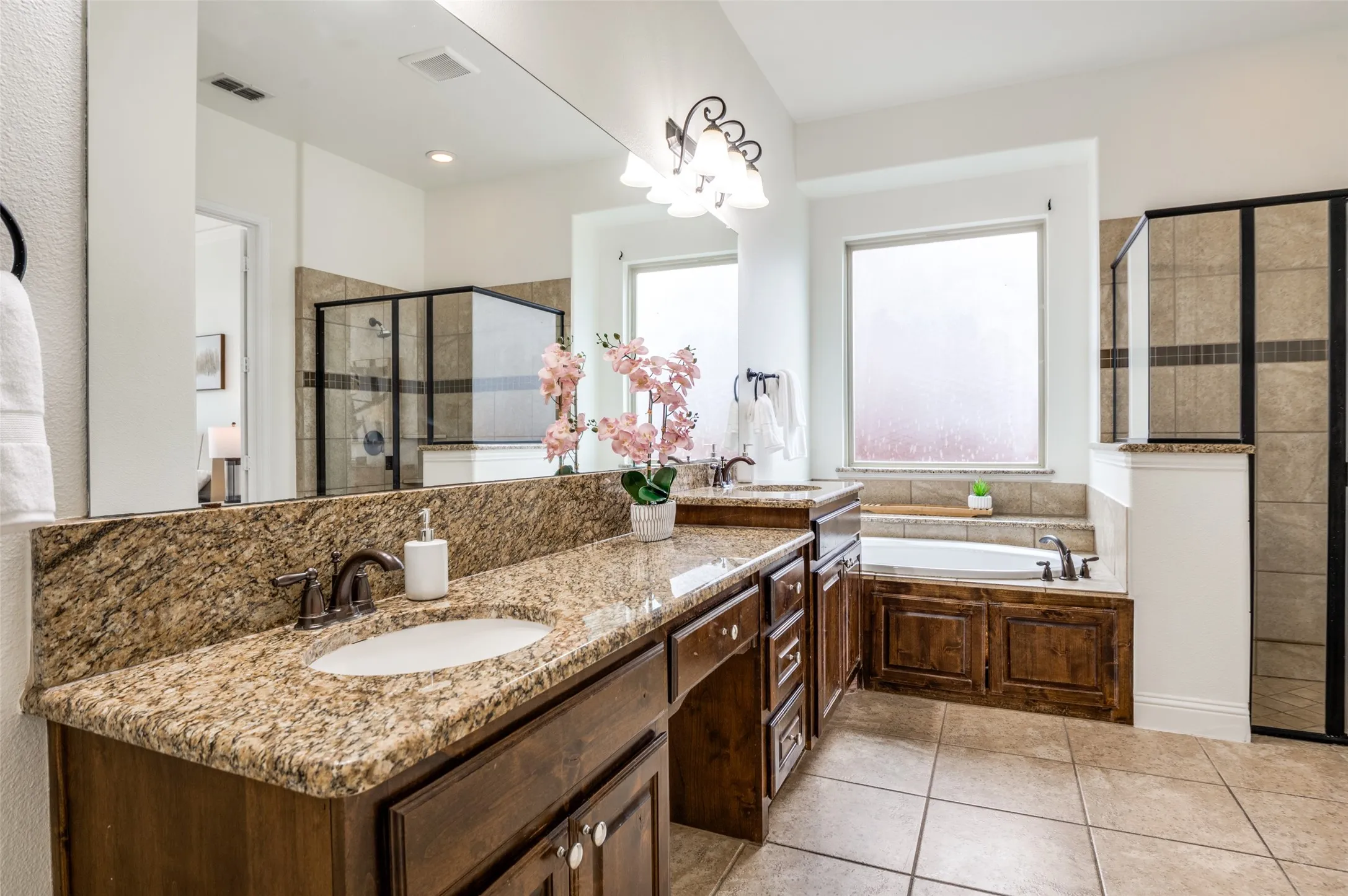 Double vanity with stone countertop, built-in soaking tub, separate glass-enclosed shower, wood-finish cabinetry, and tiled flooring