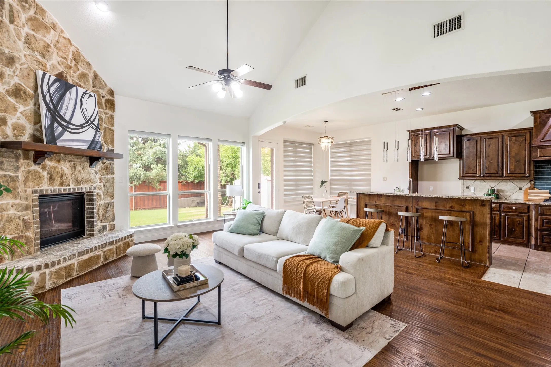 Vaulted ceiling with ceiling fan, stone fireplace with hearth and wood-finish mantel, multiple windows, wood-finish flooring, and a kitchen with wood cabinetry