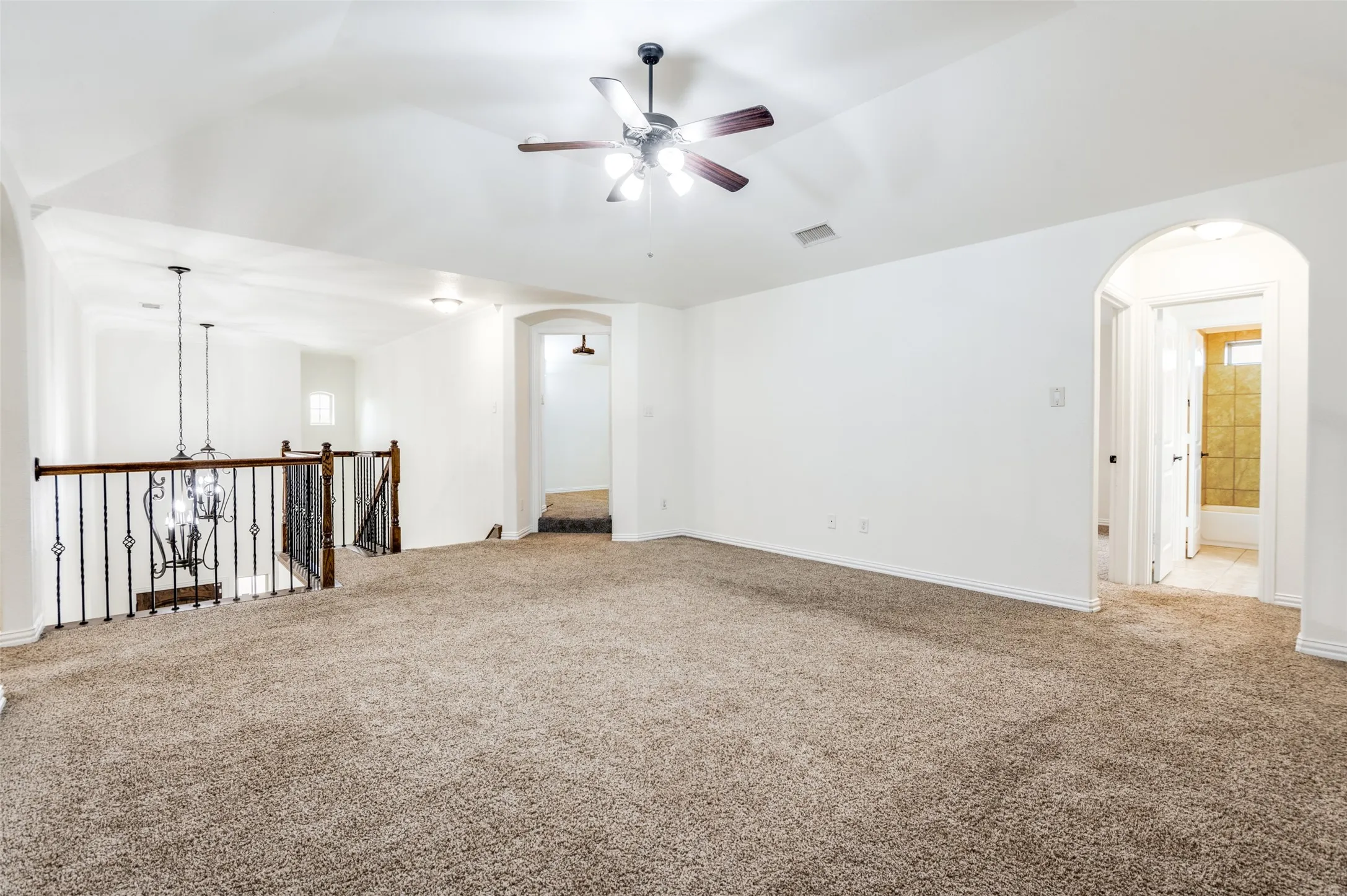 Spacious carpeted area featuring a vaulted ceiling with a ceiling fan, arched doorways, and a staircase with wrought iron balusters