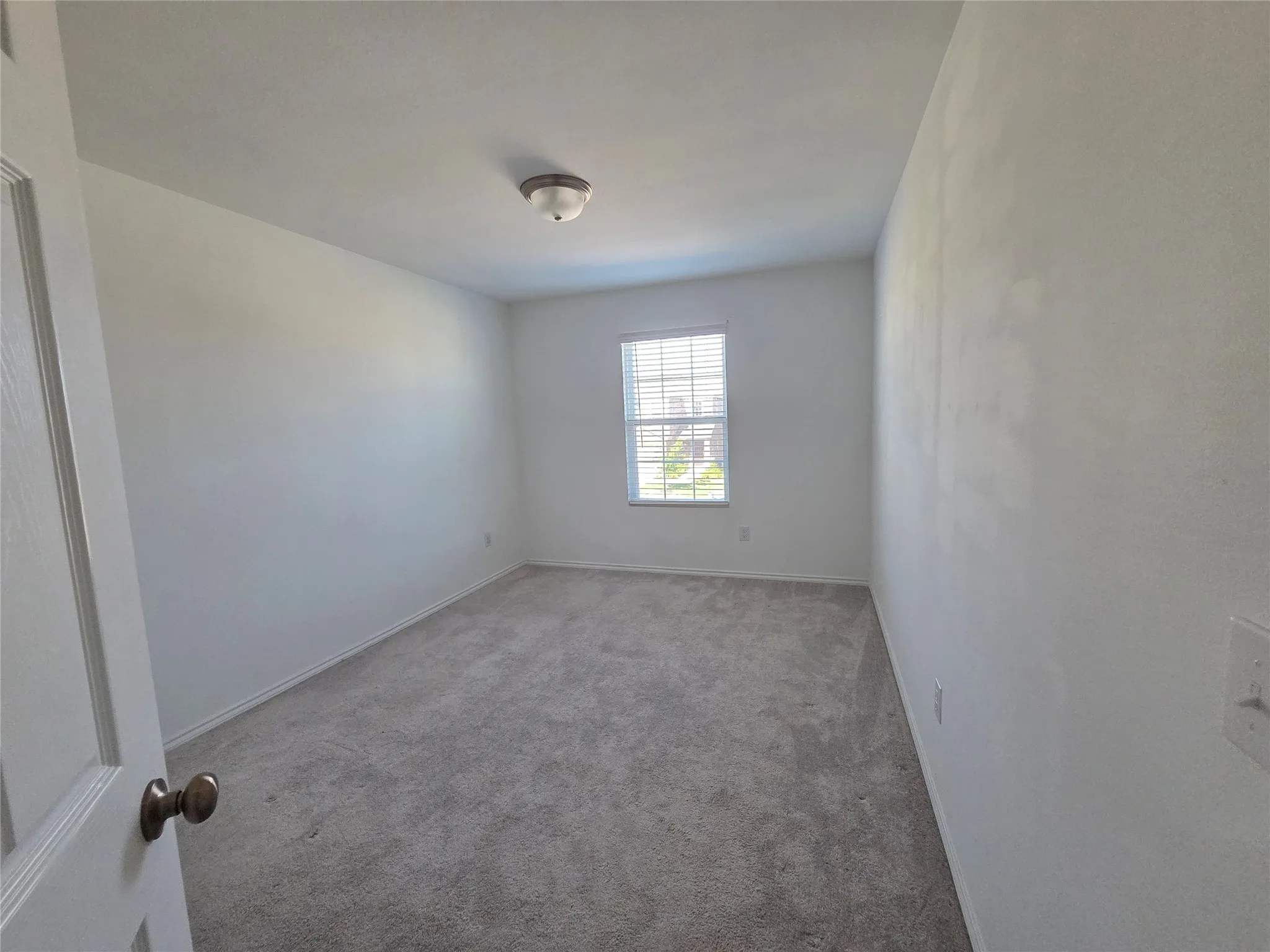 Wall-to-wall carpeting, window with blinds, ceiling-mounted light fixture, and a solid door with a bronze-finish knob