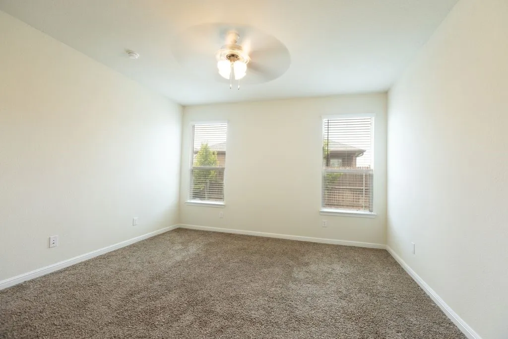 Neutral wall paint, plush carpet flooring, two windows with blinds, and a ceiling fan with integrated lighting