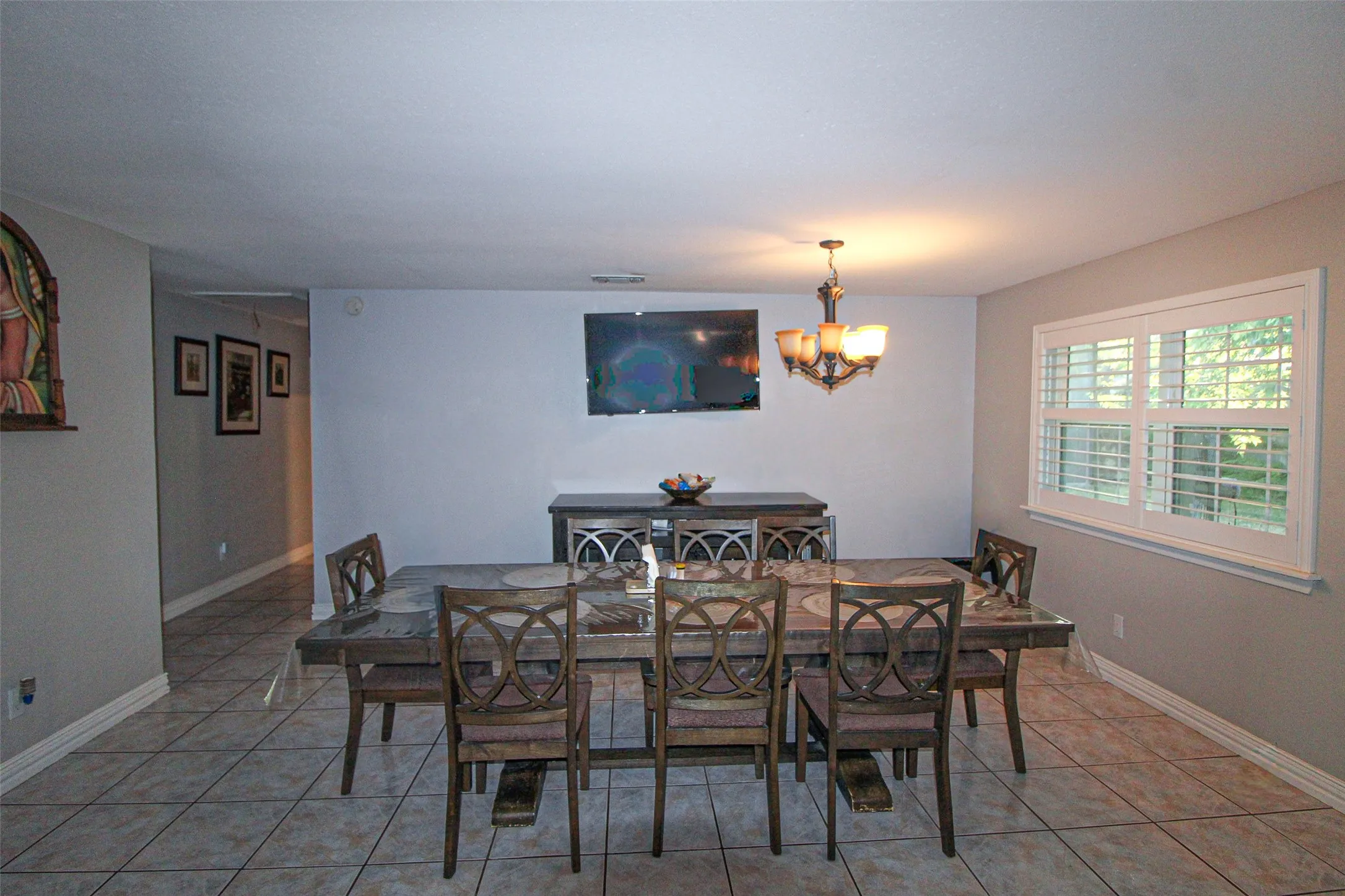 Spacious dining area featuring tile flooring, a multi-light chandelier, a large window with plantation shutters, and light-toned wall paint
