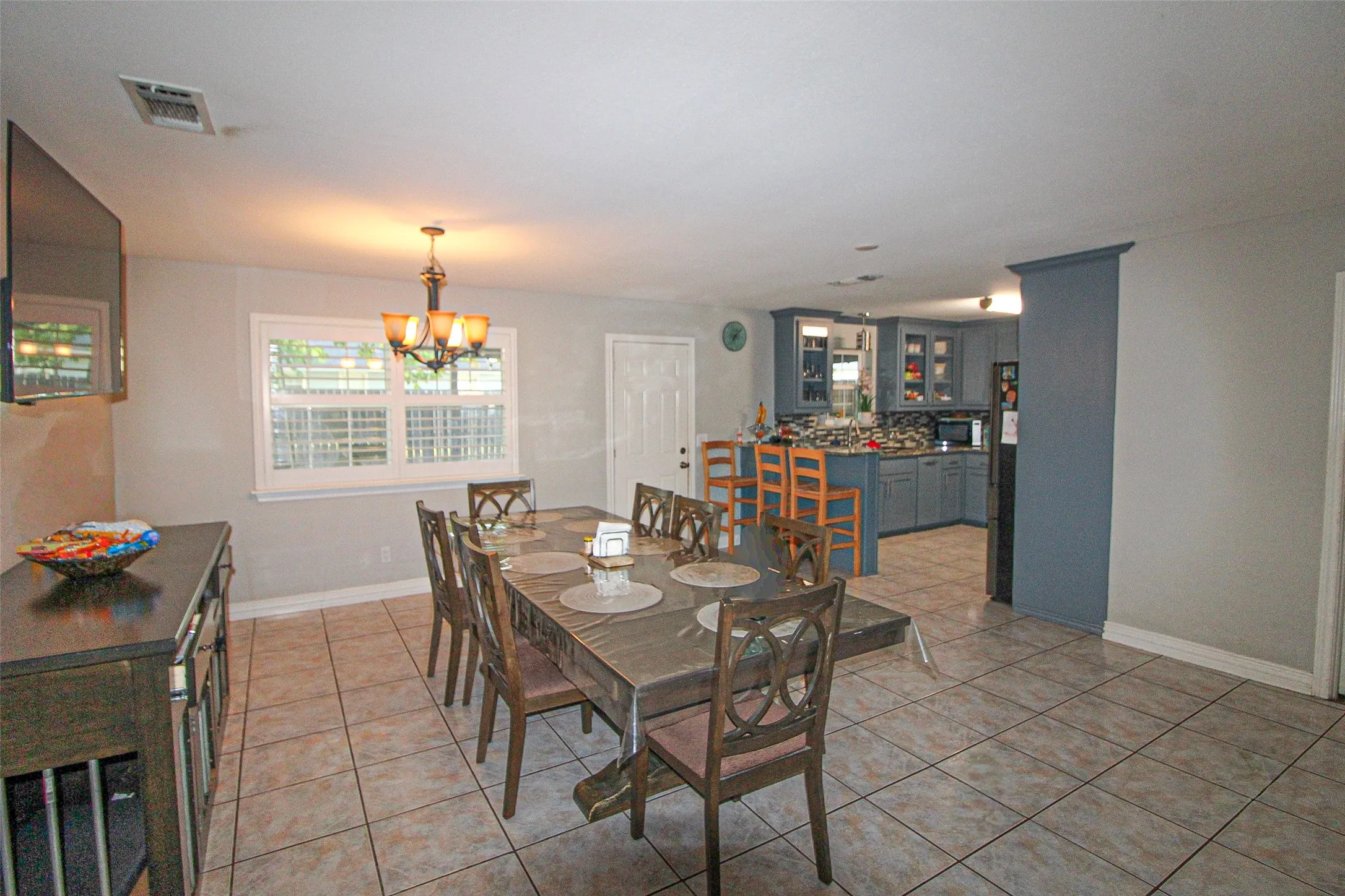 Open-concept dining area featuring tiled flooring, a chandelier light fixture, and a window with plantation shutters, with a kitchen showcasing blue cabinetry, a tile backsplash, and dark appliances