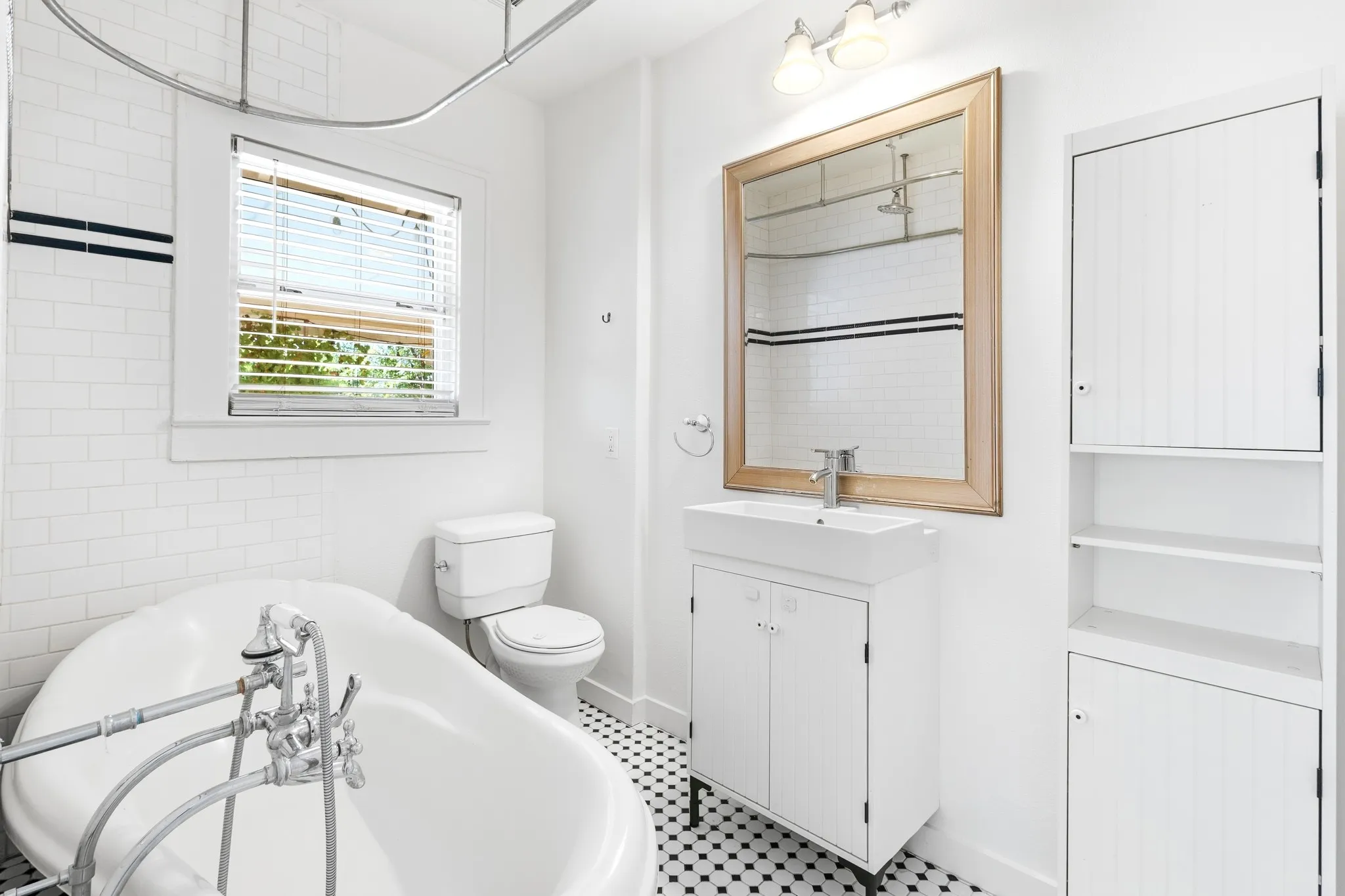 Freestanding soaking tub with chrome fixtures, white subway tile with accent banding, black and white mosaic floor tile, white vanity with integrated sink, and an adjacent built-in storage cabinet