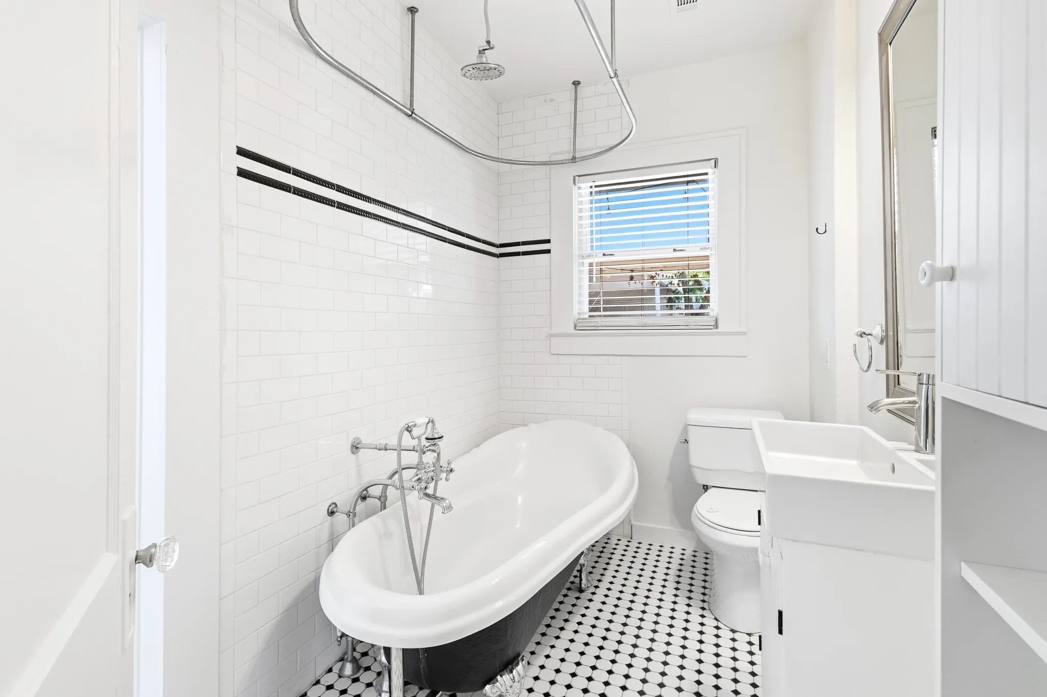 Freestanding bathtub with chrome fixtures, subway tile walls with black accent banding, black and white hexagon tile flooring, pedestal sink with chrome faucet, and a window with blinds