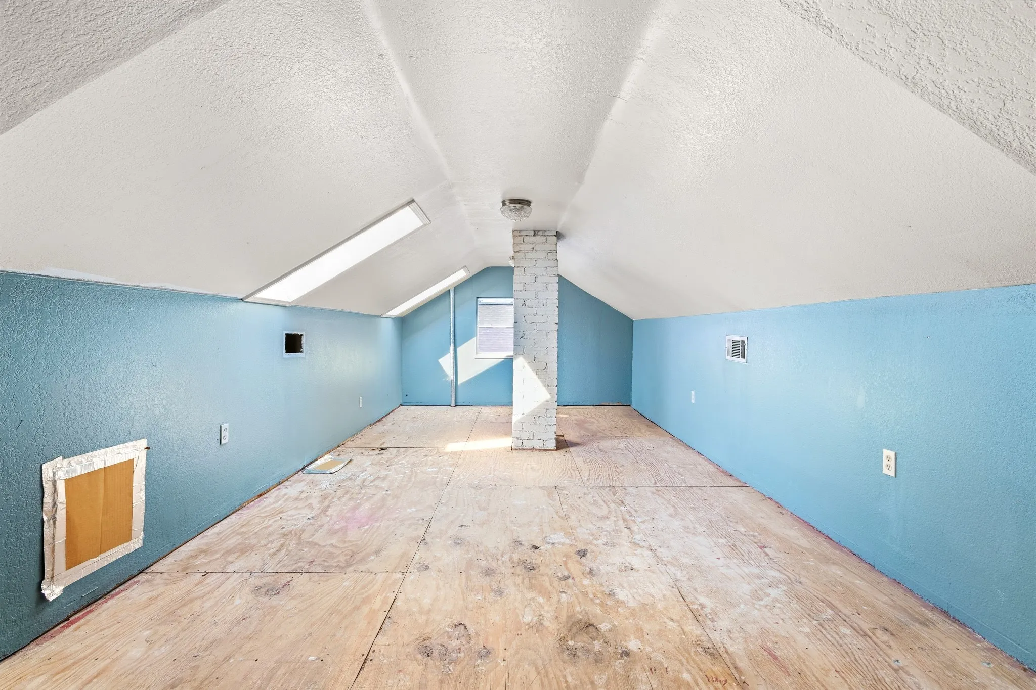 Sloping white ceilings with skylights, light blue textured walls, unfinished wood flooring, and a white brick-clad support column