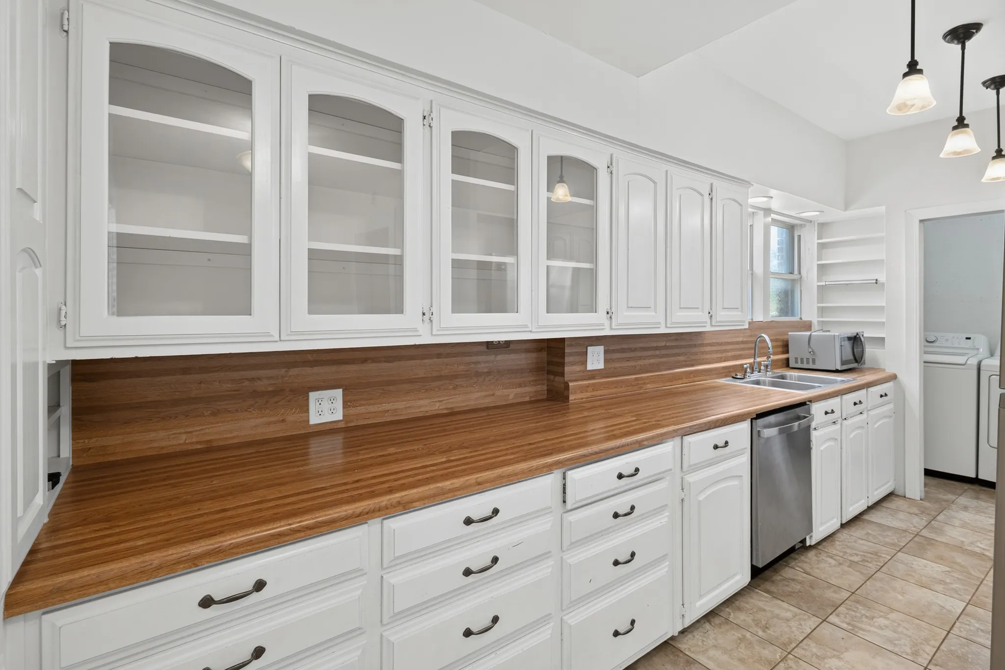 White cabinetry with glass-front uppers, wood-finish countertops, stainless steel dishwasher, and a double basin sink