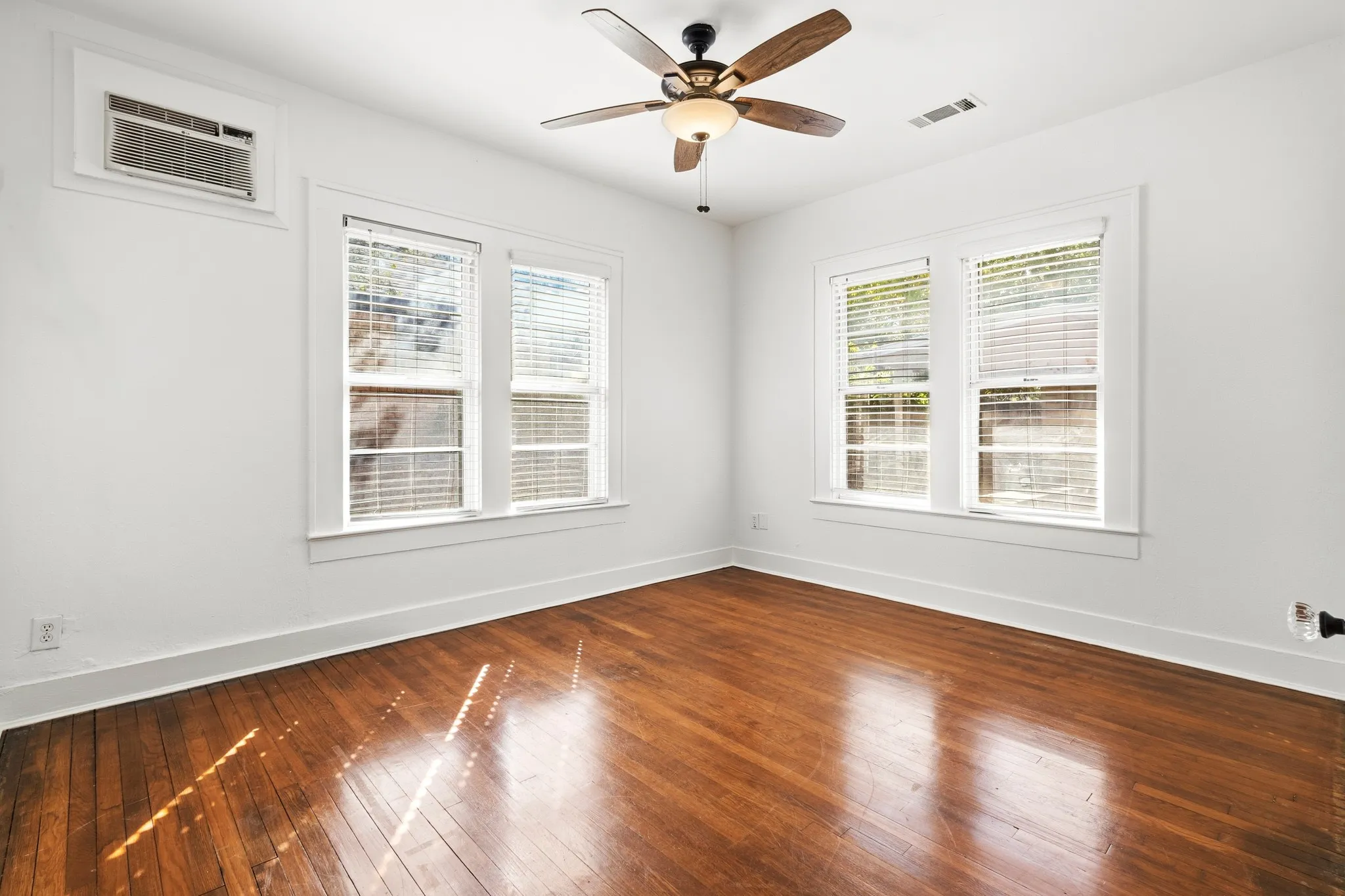 Hardwood flooring, two windows with blinds, ceiling fan with integrated lighting, window-mounted air conditioning unit