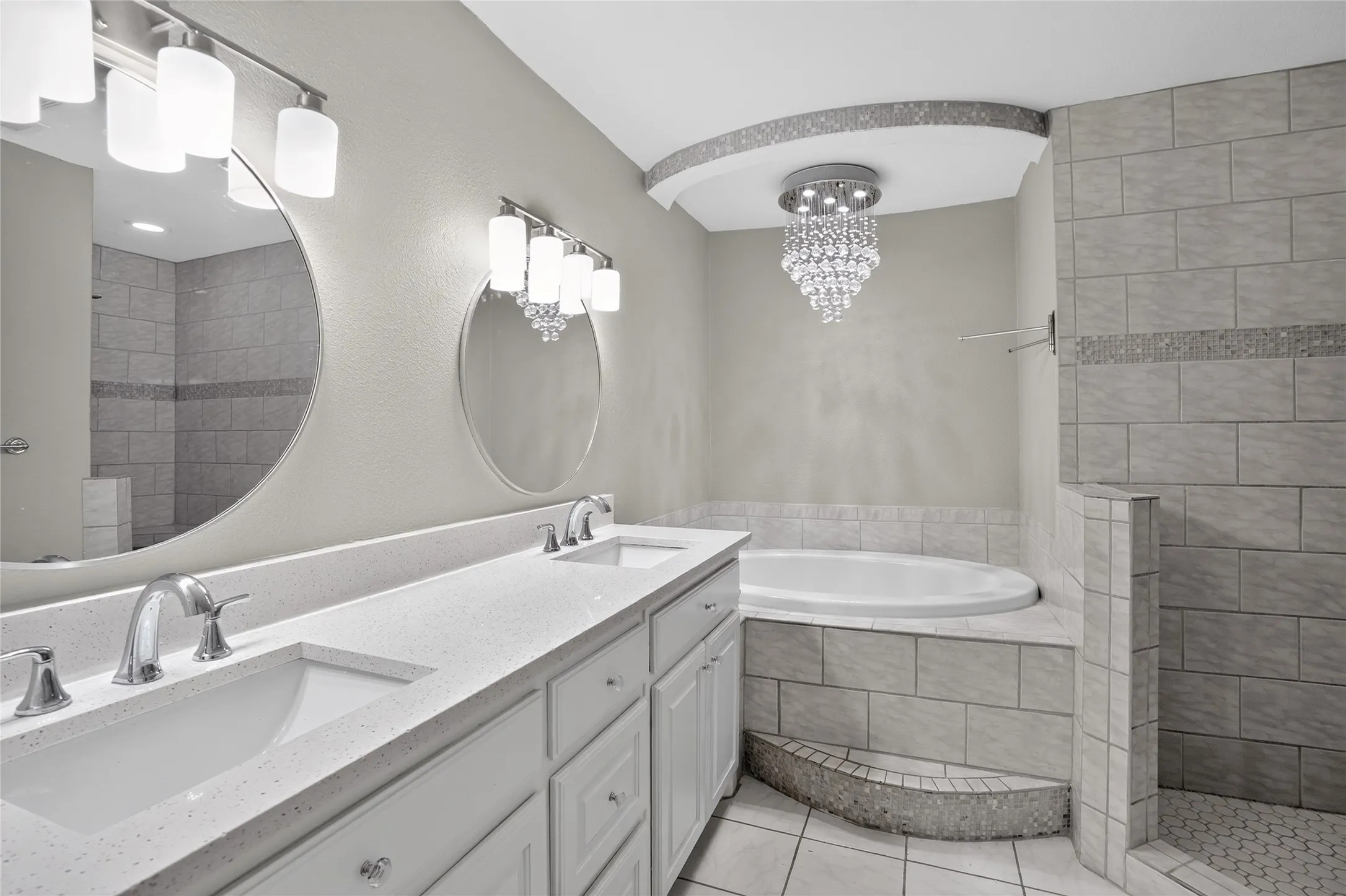 Dual vanity with integrated sinks and white countertops, built-in soaking tub, tiled shower with accent strip, and a recessed ceiling chandelier