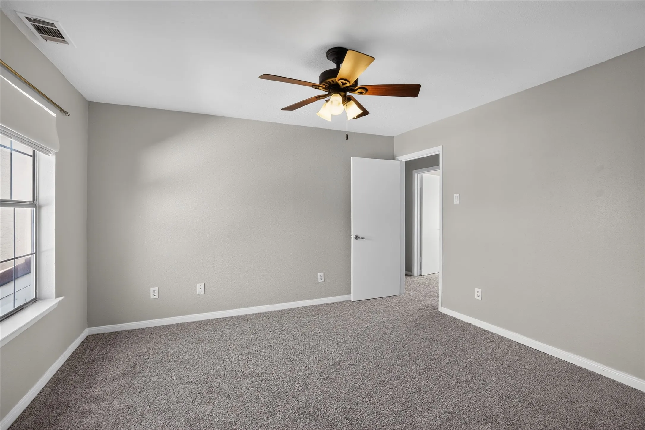 Neutral tone wall paint, textured carpet flooring, window with roller shade, ceiling fan with integrated lighting