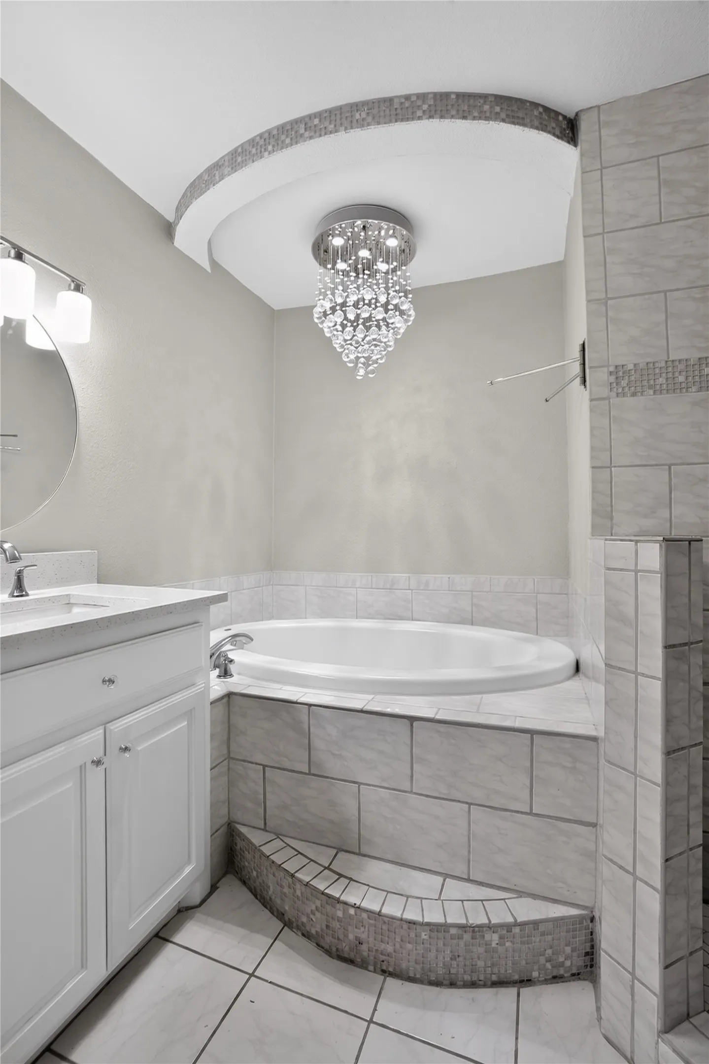 Soaking tub with tiled surround, integrated step with mosaic tile, recessed ceiling detail with mosaic trim, crystal chandelier, and white vanity with stone countertop