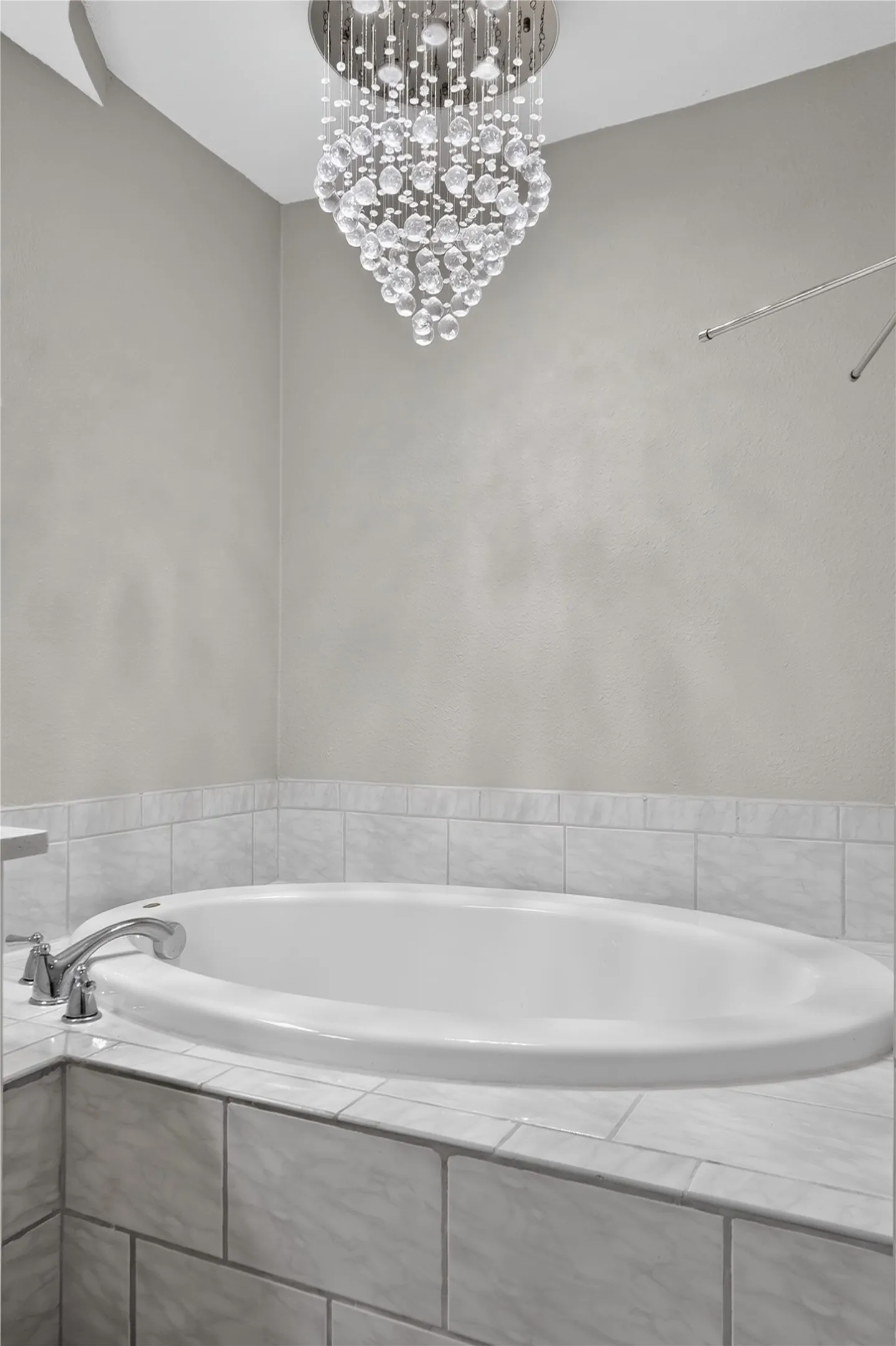 White drop-in bathtub with a chrome faucet, surrounded by tiled decking and wainscoting, illuminated by a multi-tiered crystal chandelier