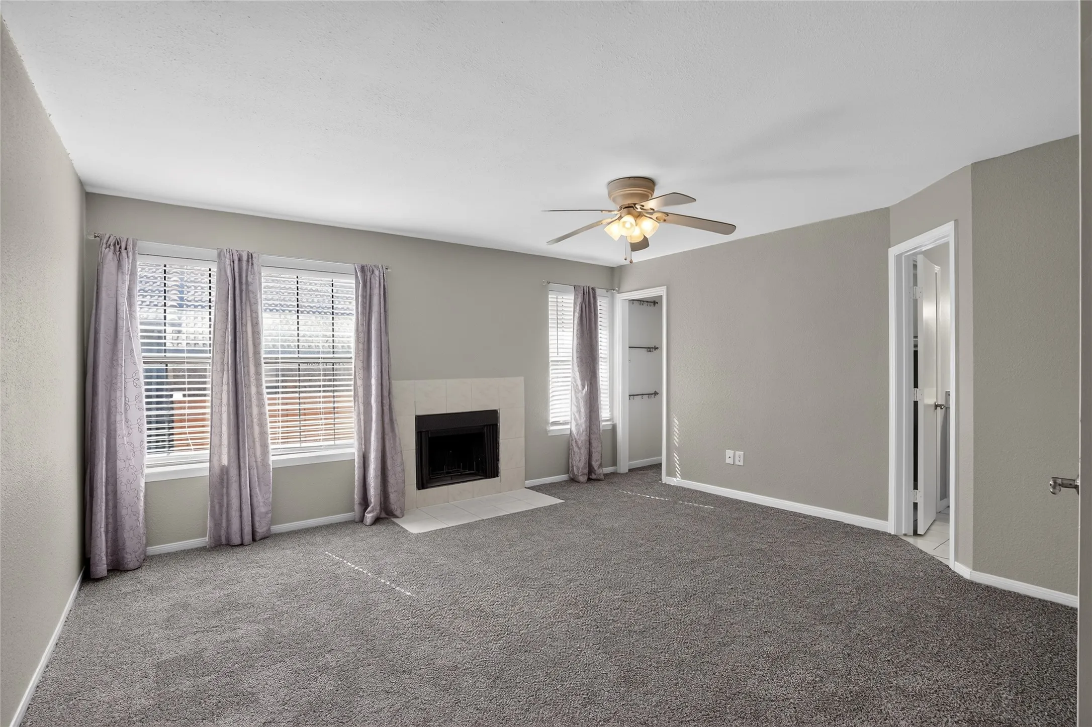 Carpeted room featuring a fireplace with a light-colored surround, multiple windows with blinds, a ceiling fan, and built-in shelving