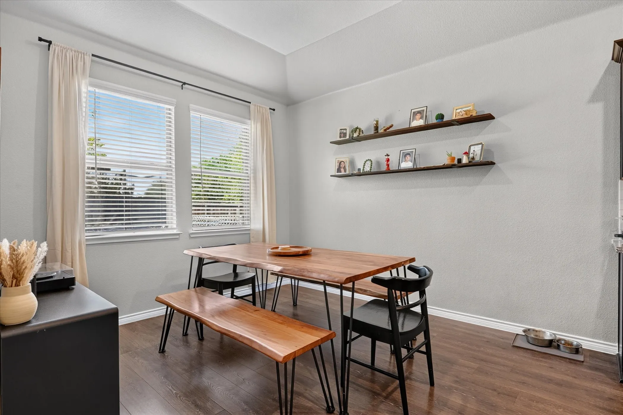 Wood-finish flooring with light gray walls, two windows featuring white blinds, and two floating shelves
