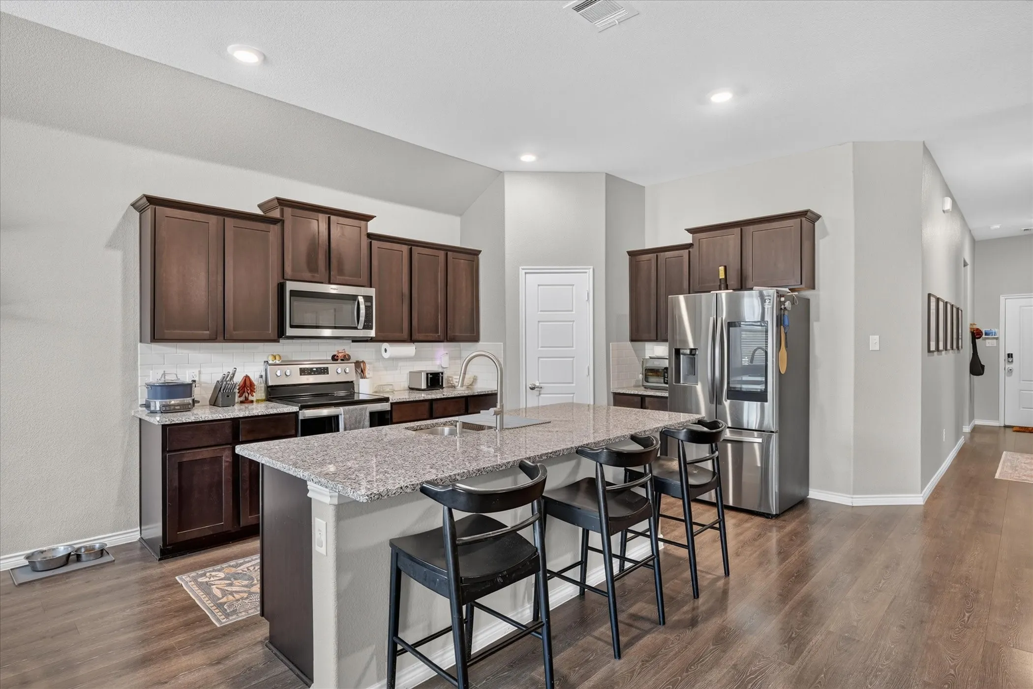 Kitchen featuring wood-finish flooring, dark wood cabinetry, stainless steel appliances, a white tile backsplash, and a large island with a light-colored countertop