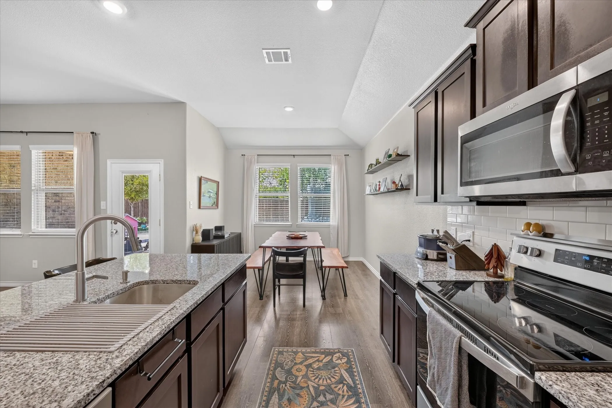 Kitchen featuring wood-finish flooring, dark wood cabinetry, light speckled countertops, stainless steel appliances, and a subway tile backsplash