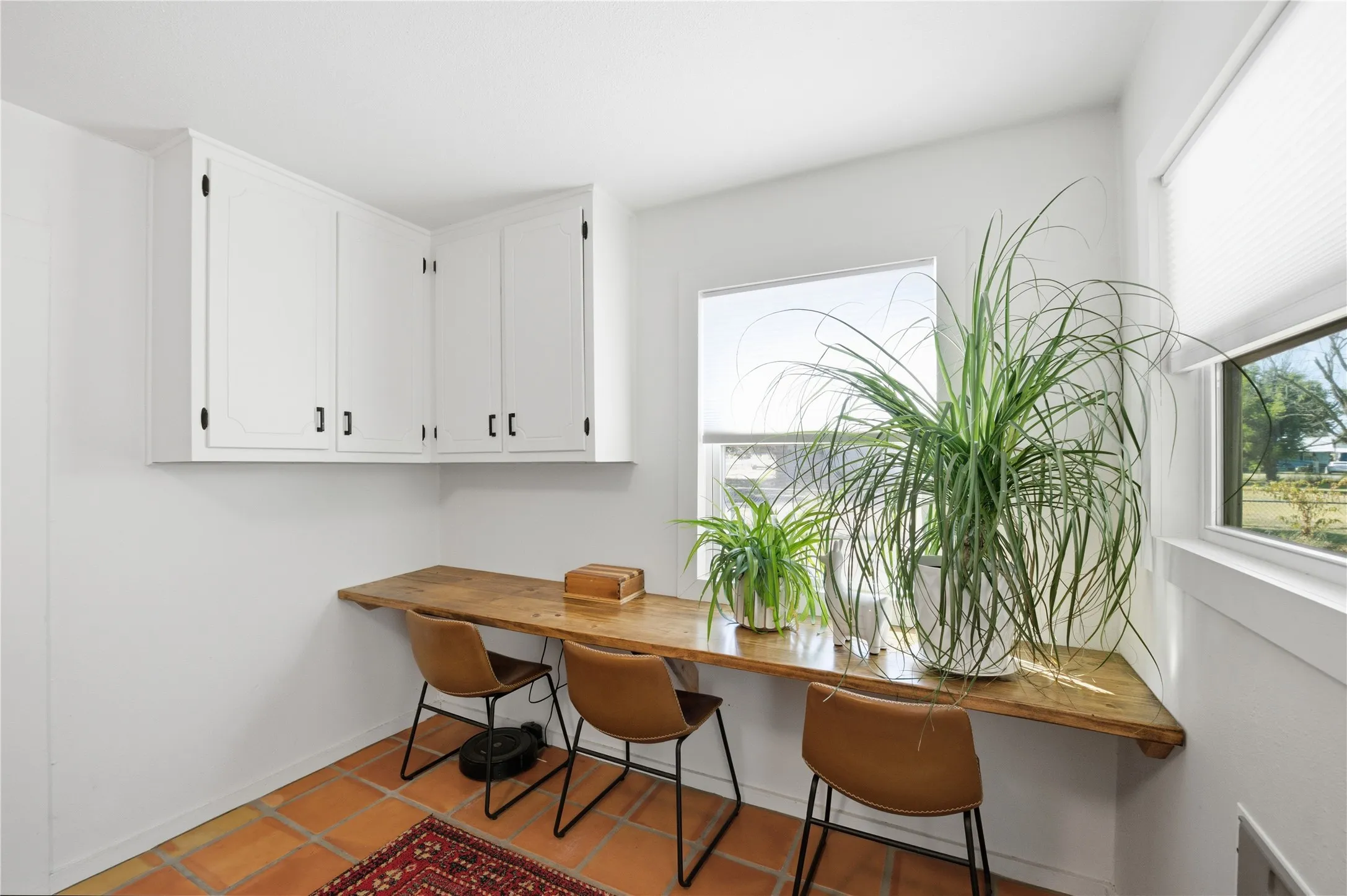 White upper cabinetry with matte black hardware, wood-finish floating desk, terracotta tile flooring, and multiple windows with cellular shades
