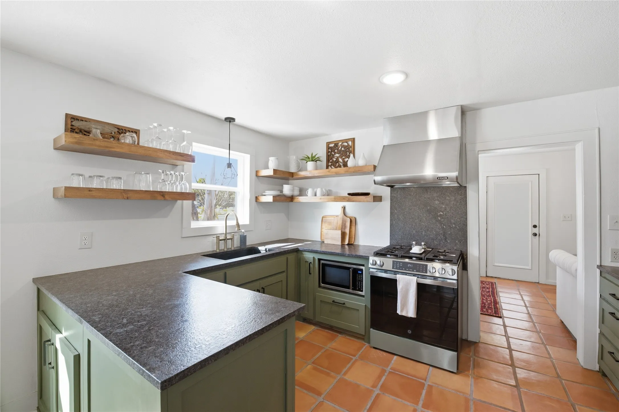 U-shaped kitchen featuring wood-finish open shelving, dark countertops, a stainless steel range with matching vent hood, and terracotta-style floor tiling