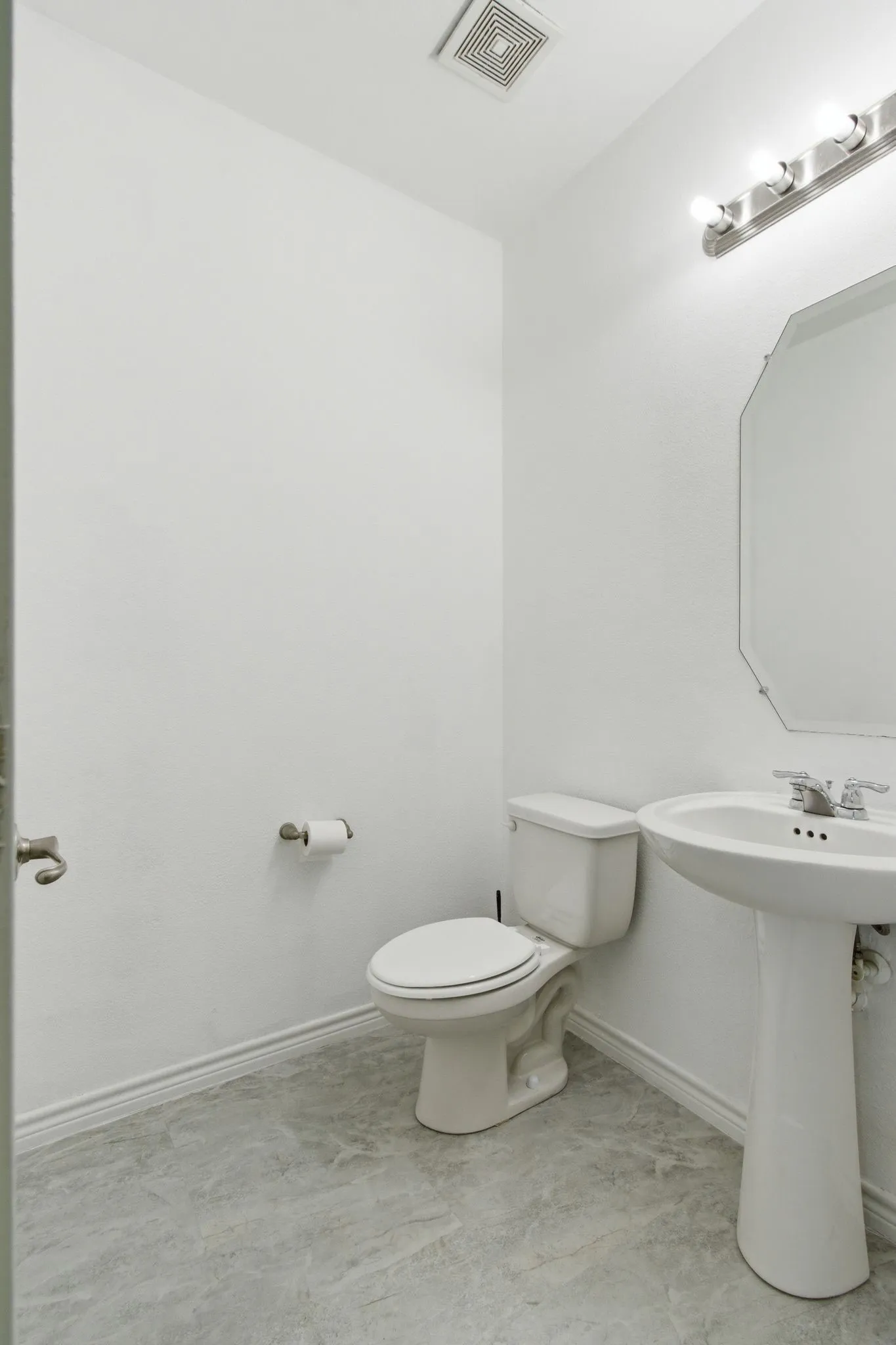 Pedestal sink with chrome faucet, octagonal mirror, wall-mounted vanity lighting, and gray-toned flooring