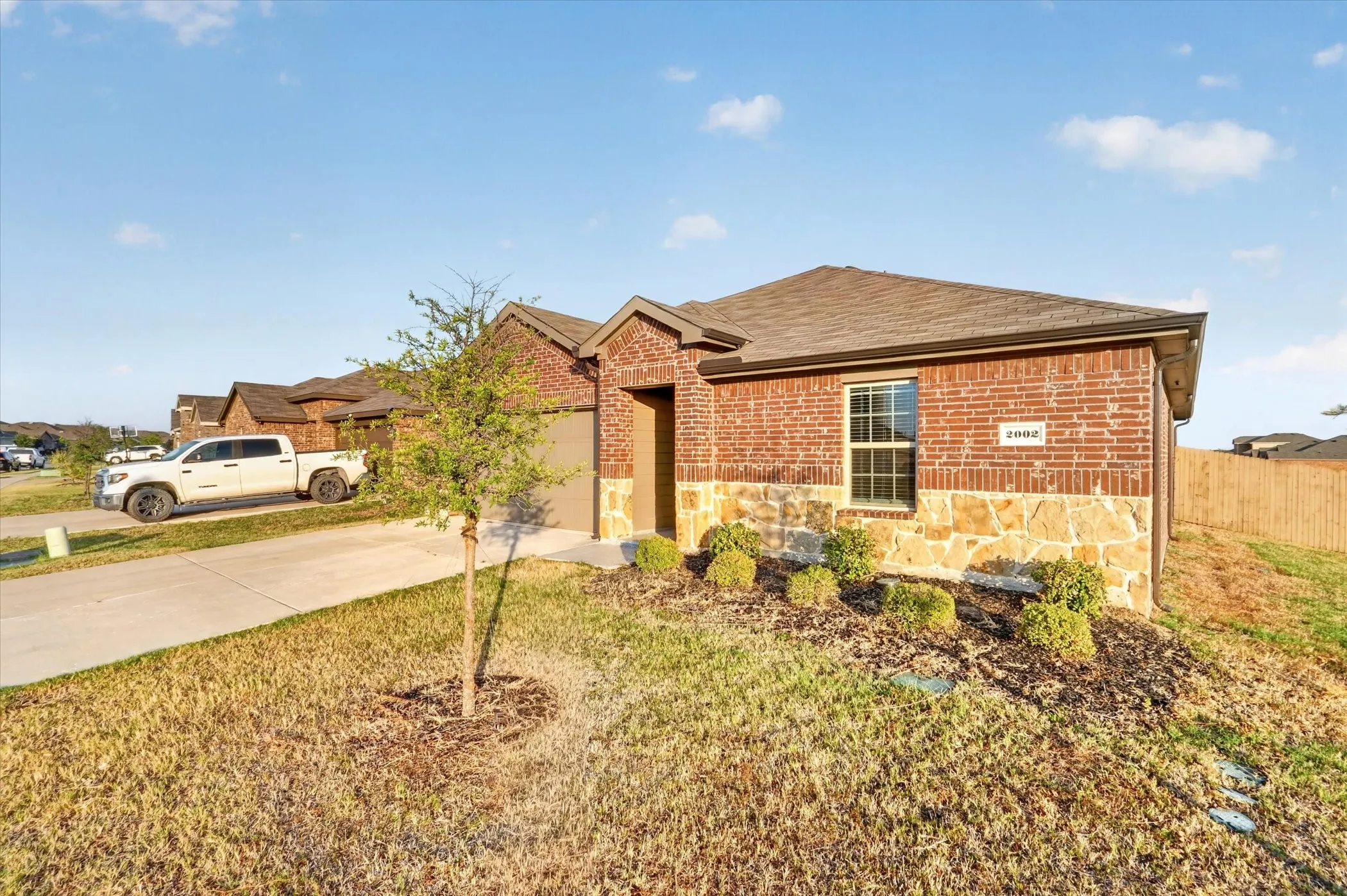 Brick and stone exterior, attached garage, well-maintained landscaping, concrete driveway, and a fenced property boundary
