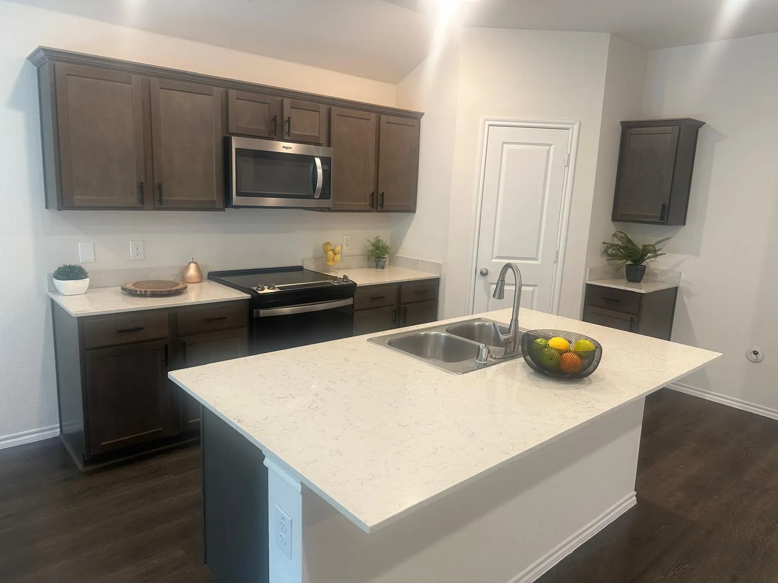 Kitchen featuring an island with a double basin sink, dark wood-finish cabinetry, light-toned countertops, and stainless steel appliances