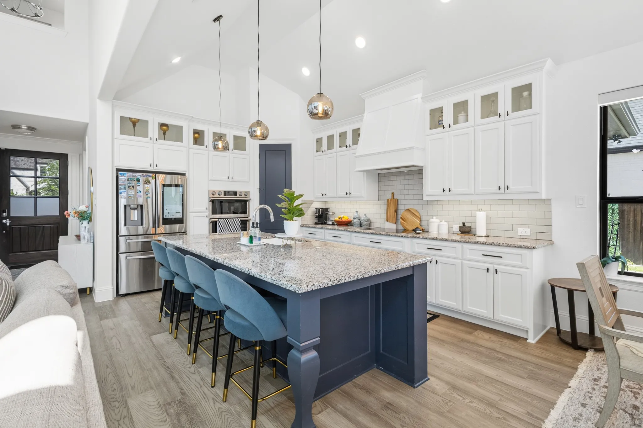Vaulted ceiling kitchen featuring a large central island with seating, granite countertops, white cabinetry with glass-front uppers, and stainless steel appliances