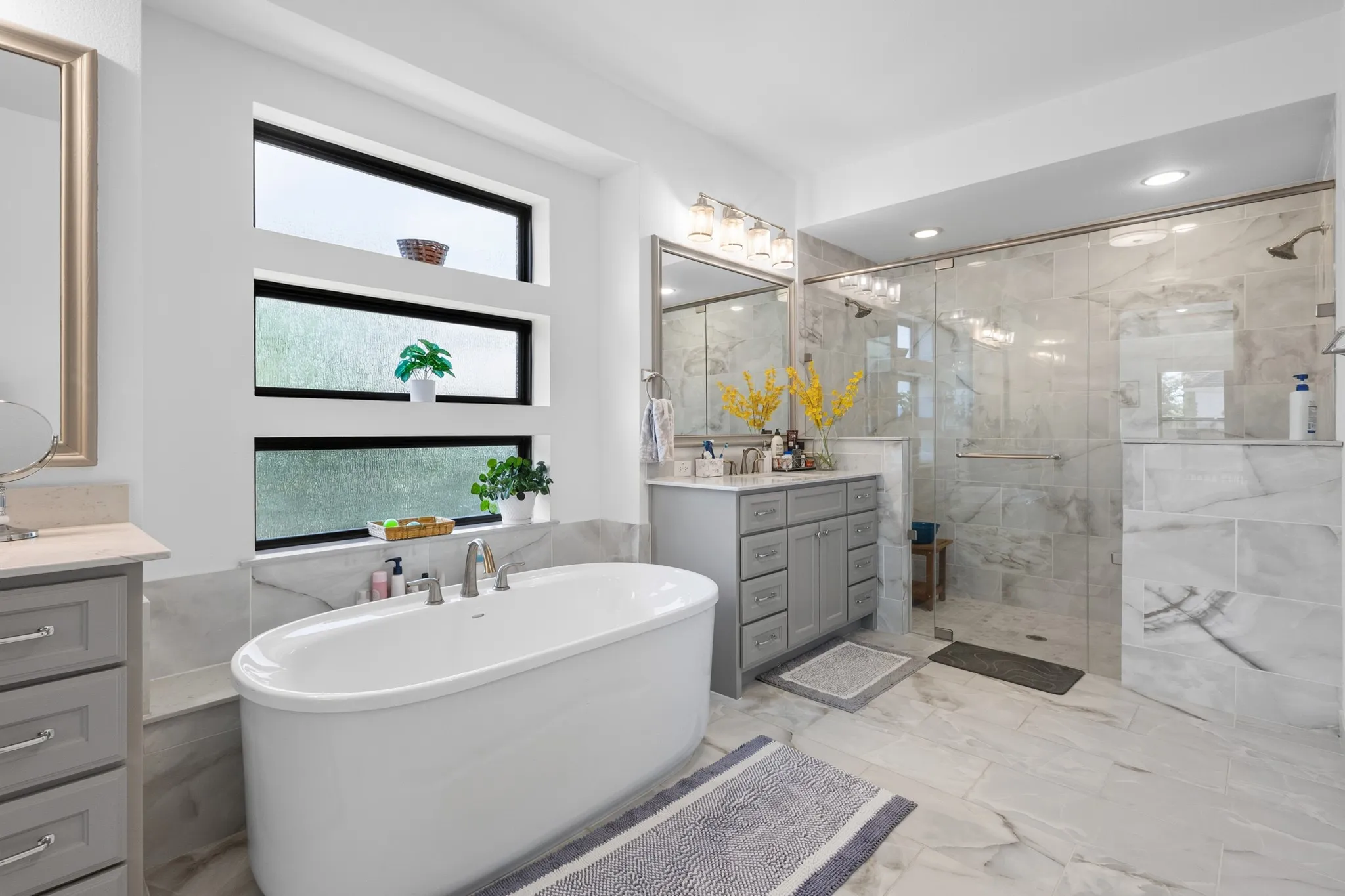 Freestanding soaking tub with a tiled surround, dual vanity with a quartz countertop, glass-enclosed shower with a rainfall showerhead, and large format tile flooring