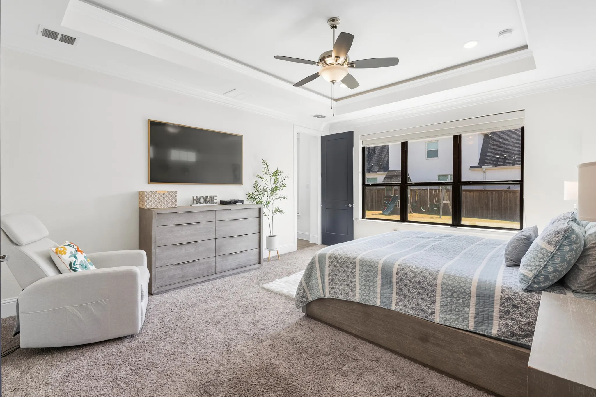 Tray ceiling with crown molding, ceiling fan with integrated lighting, large picture window, and dark wood-finish door