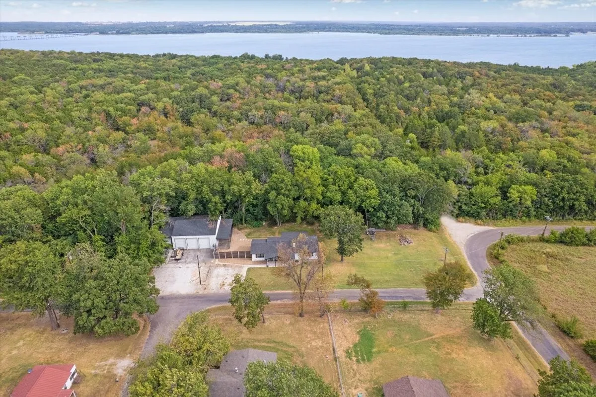 Aerial view showcasing a residential property nestled within mature hardwood forest, featuring a modern structure with clean architectural lines, expansive manicured grounds, paved driveway approach, and panoramic water vistas across the tree canopy in the distance