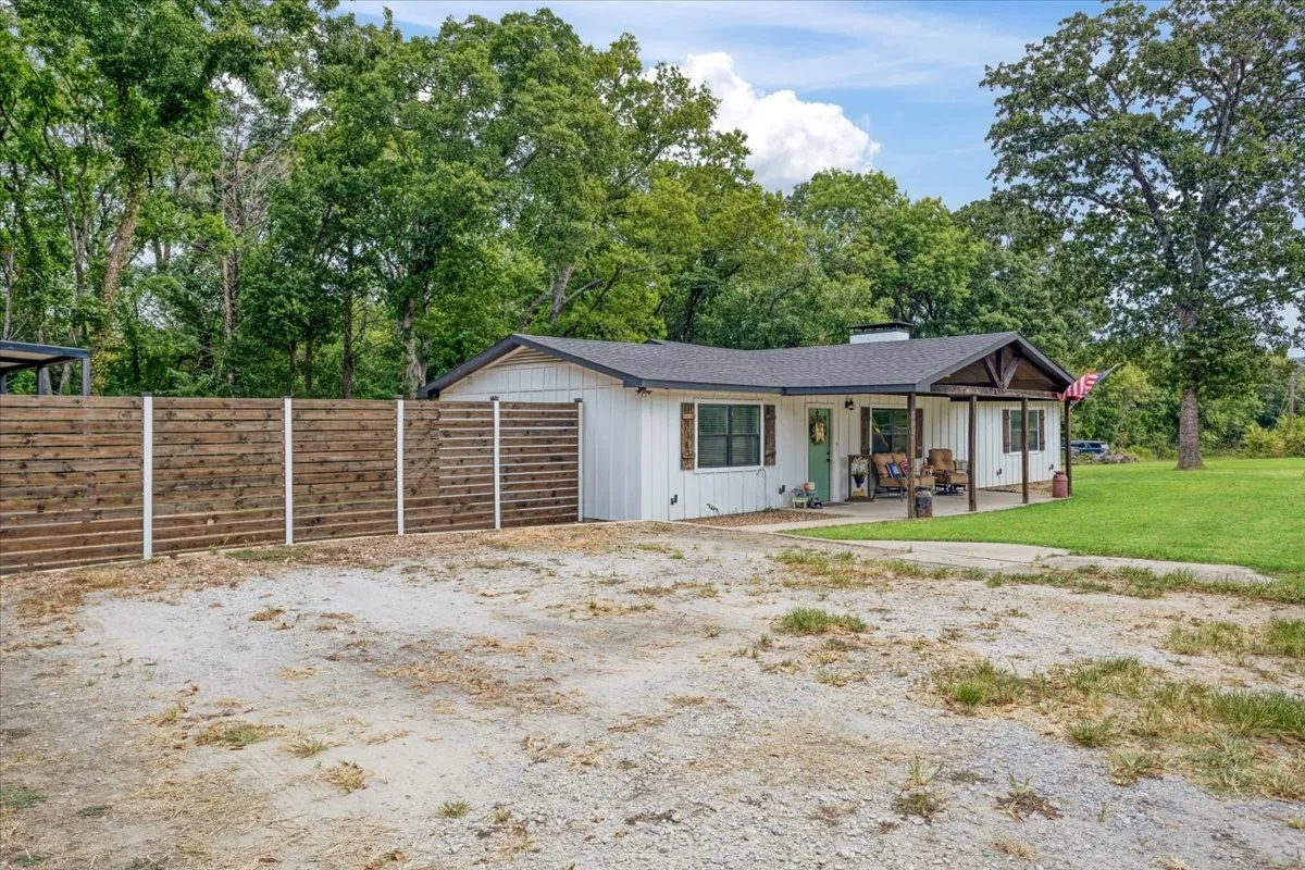 White exterior with dark trim and shutters, covered front porch, horizontal wood privacy fence, gravel driveway, and surrounding mature trees