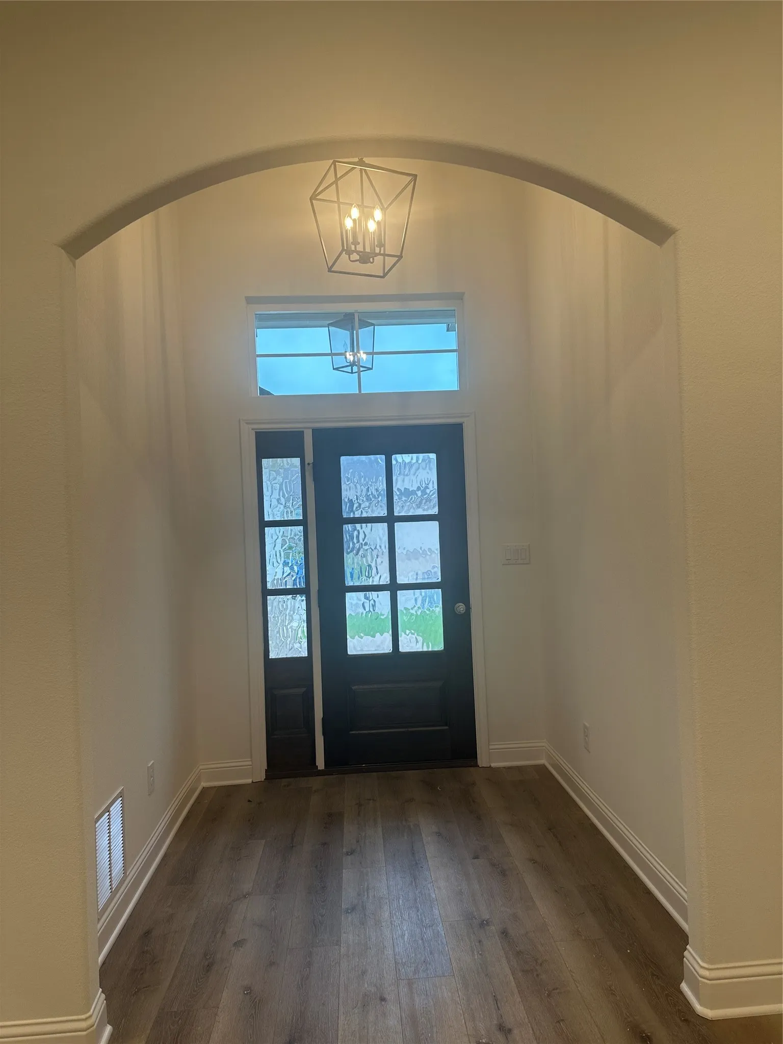 Archway entry featuring wood-finish flooring, a contemporary geometric pendant light, and a dark-paneled door with decorative glass inserts and a transom window